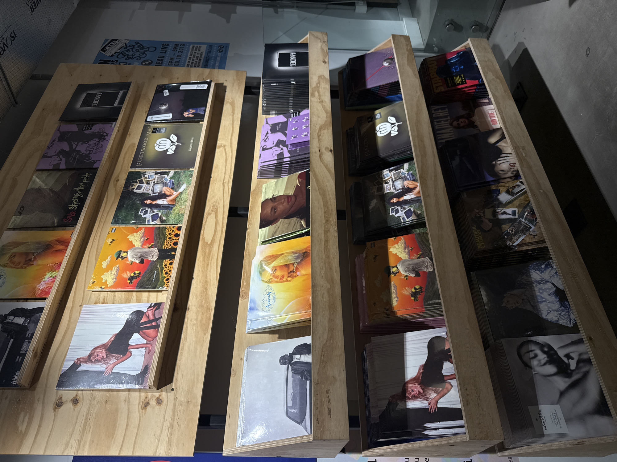 The image shows a display of vinyl records in a store. The records are arranged in wooden shelves, angled slightly for better viewing. Album covers are visible, with artists like THE 1975, Fleetwood Mac, and Lana Del Rey featured. The display is set against a backdrop that includes a poster indicating a "LEAU LIVE" event at "OUTFITTERSLER 33" on "09.10.2013". The presence of this poster hints at the location being potentially associated with OUTFITTERSLER 33, although the exact city is not stated in the given text. There are no people visible in the image. The environment appears to be a well-lit, modern retail space, with details like ceiling fixtures and wall panels adding to the context.