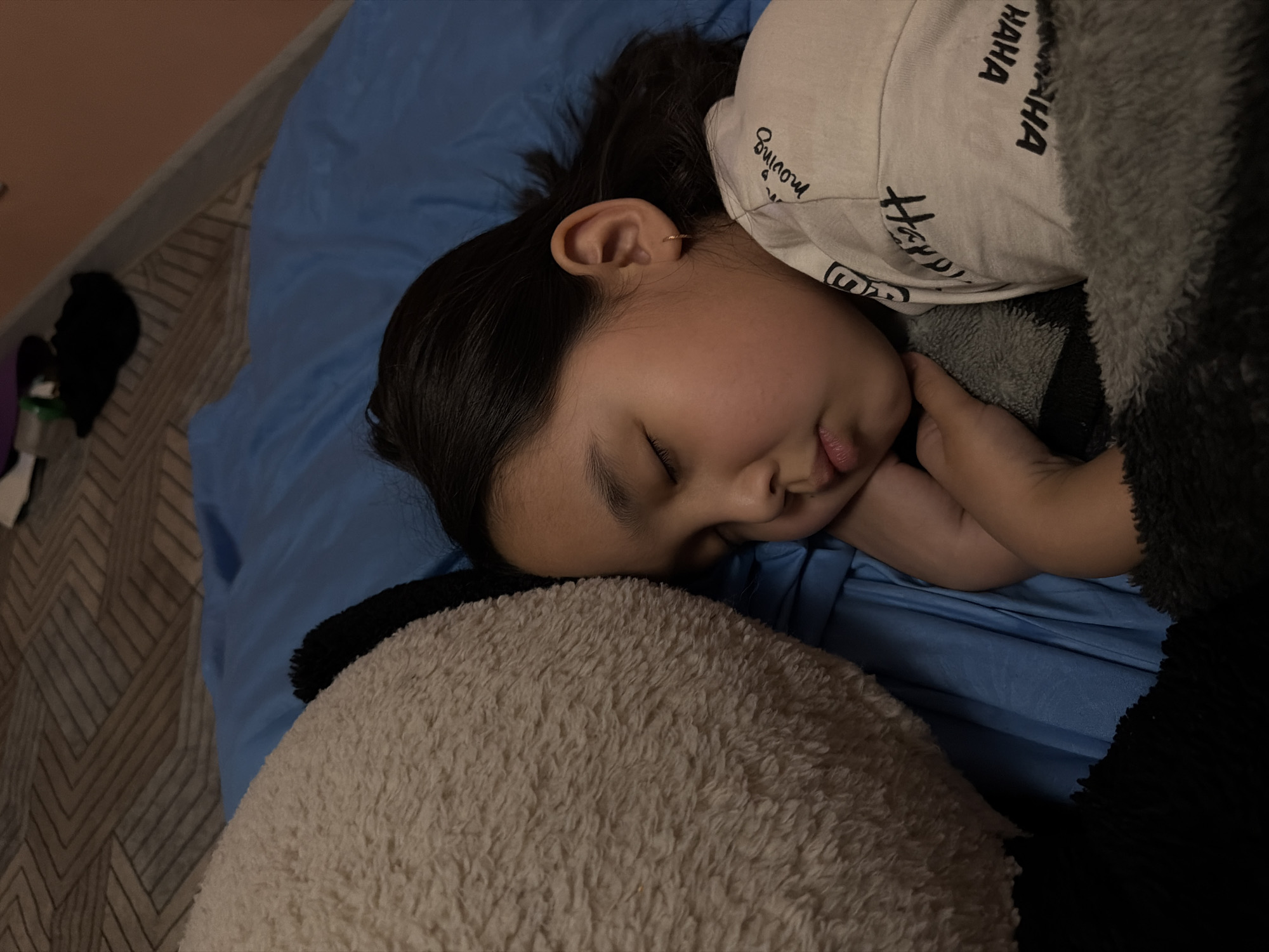 An overhead photograph captures a young person, likely a child, sleeping on their right side in a bed in Unknown, Russia. The individual has dark hair and a light complexion, with their eyes closed in slumber. A small, golden hoop earring is visible in their right ear. They are wearing a light-colored, long-sleeved shirt featuring black text, including the words "HA HA HA" and "moving" partially visible. Their left hand is tucked under their chin/cheek area. To the left of the sleeping individual is a large, off-white/beige, shaggy plush object, possibly a stuffed animal, which the person appears to be resting against or cuddling. The bed is covered with a plain blue sheet or duvet. A grey and black patterned, fluffy blanket or garment covers a portion of the person's lower body and the plush toy. In the upper background, a patterned bedspread or blanket with geometric designs in brown and grey tones is visible. Further in the upper right, a peach-colored wall with a white baseboard is present, and some indistinct dark objects or clutter can be seen on a surface. The overall lighting suggests an indoor setting, possibly a bedroom.