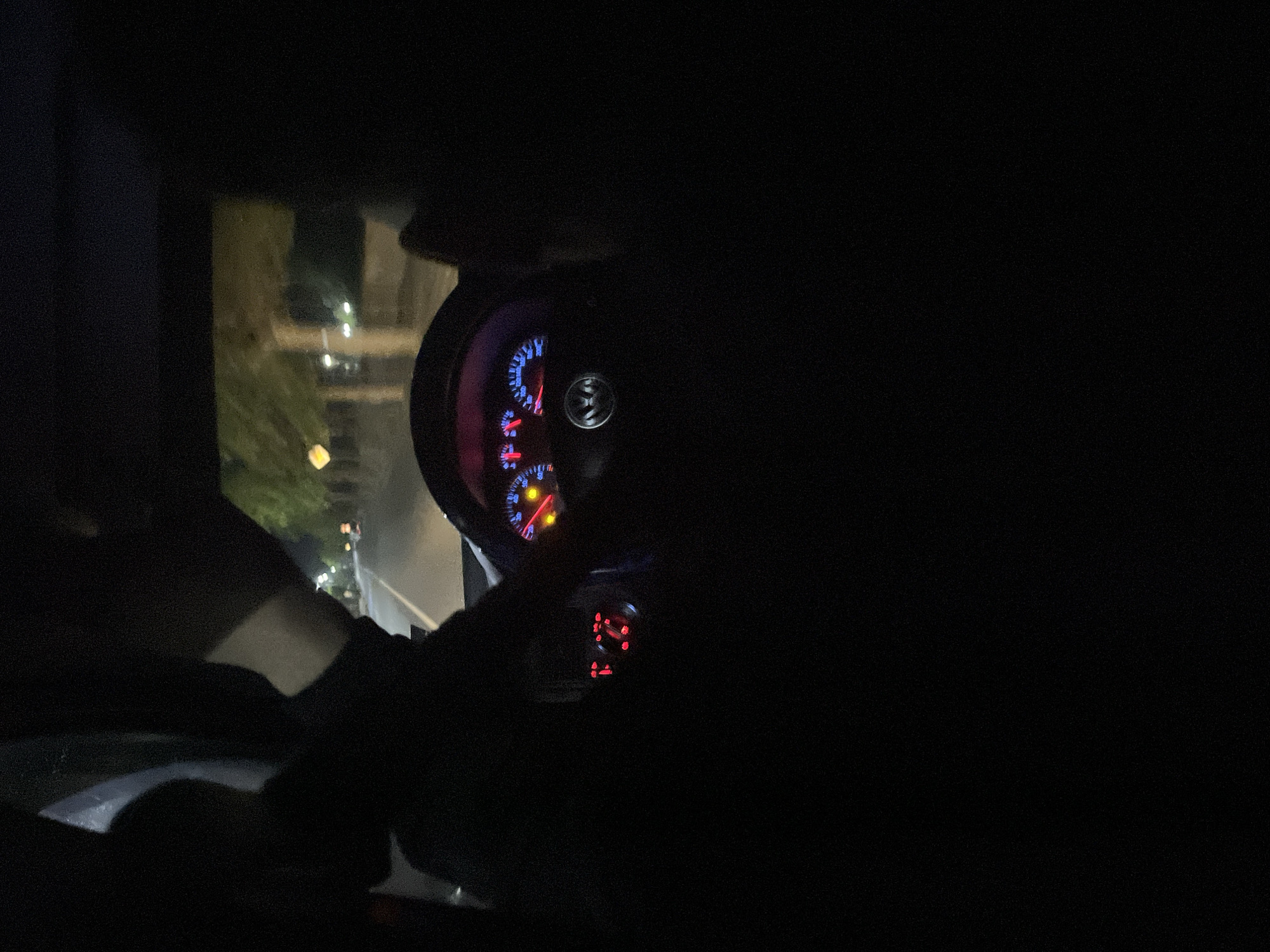 The image depicts the interior of a vehicle, likely at night. The illuminated dashboard is visible, showing a speedometer with blue and red markings. The steering wheel is partially visible on the left, and parts of an arm and sleeve are visible. Outside the window, blurry streetlights and buildings suggest an urban setting. Given the information provided, the photo was taken inside the vehicle in the city of Zlatograd, Bulgaria.