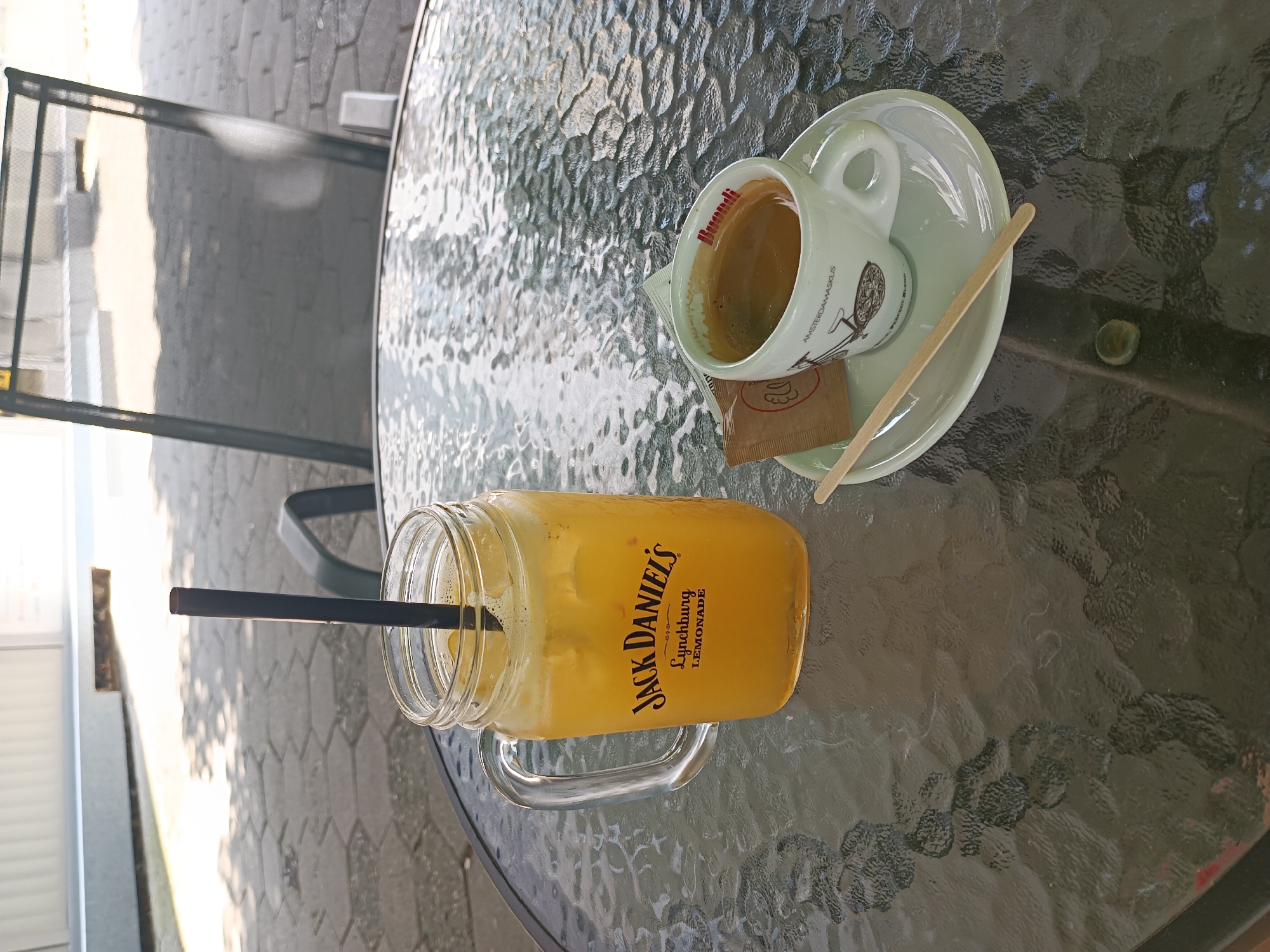 A close-up, eye-level shot depicts a glass table on an outdoor patio, likely at a cafe in Burgas, Bulgaria, bathed in natural sunlight. The table's textured glass surface reflects the surroundings. On the table, a clear glass jar with a handle contains a yellow, iced beverage with a black straw. The jar is branded with "Jack Daniel's Lynchburg Lemonade." To its right, a small white coffee cup with a saucer holds a dark espresso. The cup is decorated with a bicycle graphic and the word "Buondi". A small brown sugar packet and a wooden stir stick lie next to the coffee cup. In the background, the blurry shapes of an outdoor chair and a paved walkway with hexagonal stones are visible, suggesting a public space. The overall scene conveys a relaxed, outdoor cafe atmosphere.