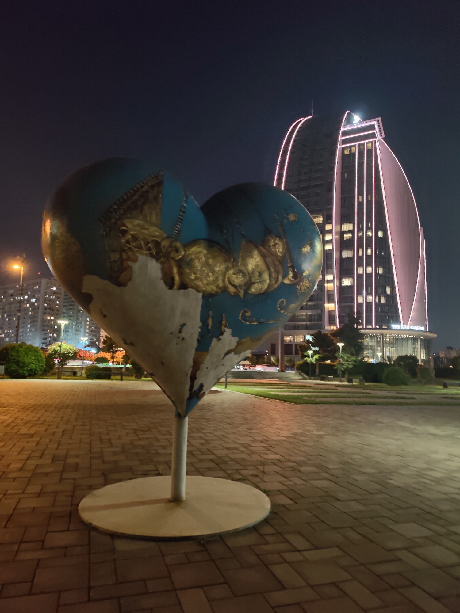 In Baku, Azerbaijan, at night, a large, blue, heart-shaped sculpture with gold embellishments stands in the foreground, mounted on a white pole and a circular base. The sculpture shows signs of wear, with chipped paint revealing a white underlayer. Behind it, a modern, curved skyscraper, illuminated by pink vertical lights, dominates the background. The building displays "THE RITZ-CARLTON" signage. The scene is set in a paved plaza, with grass, trees, and benches in the middle ground. In the distance, apartment buildings are visible, lit by numerous windows and streetlights. The sky is dark, indicating nighttime.