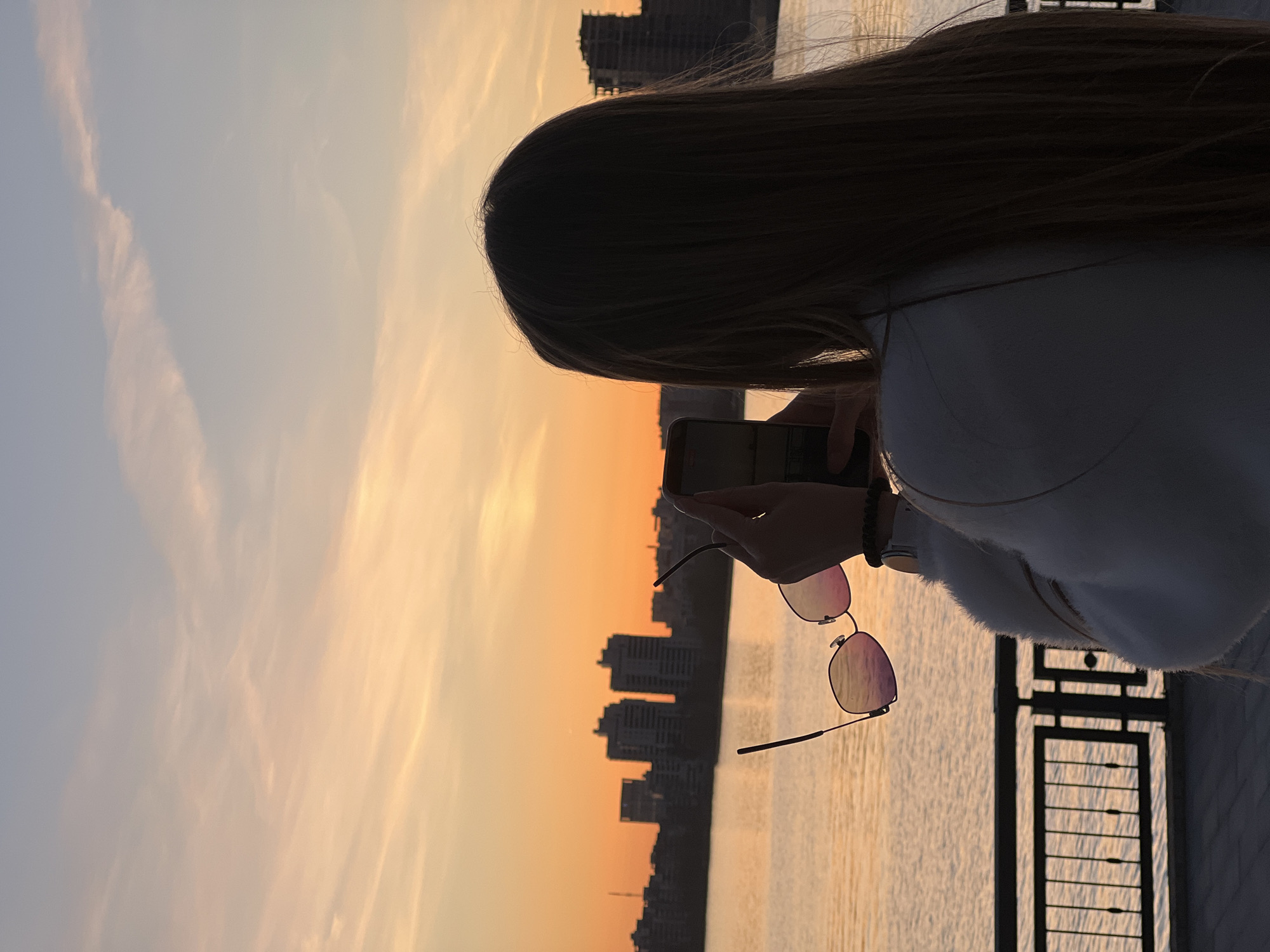 The image captures a serene sunset scene in Kazan, Russia, featuring a person from a rear perspective. The central subject is a person with long, dark hair, seen from the shoulders up, facing away from the viewer. They are holding a smartphone in their right hand, possibly taking a photo or video, or browsing. A pair of sunglasses with reflective, pinkish lenses is held by its frame, dangling from the person's fingers.
The setting is an outdoor embankment or promenade, suggested by the dark metal railing visible in the lower right corner, separating the viewer from a wide body of water. The water reflects the warm, golden light of the setting sun, appearing calm with gentle ripples.
Above the water and behind the person, a striking sunset dominates the sky. The sky transitions from a soft, light blue and gray at the top to vibrant shades of orange, yellow, and pink closer to the horizon. Faint, wispy clouds are scattered across the colorful sky. A silhouette of a city skyline, comprising multiple buildings of varying heights, is visible across the water on the far bank, framed by the sunset glow.
The time of day is clearly sunset, as indicated by the rich, warm colors in the sky and the silhouetted elements. The overall atmosphere is tranquil and contemplative, suggesting an appreciation of the natural beauty. No discernible text is visible in the image.