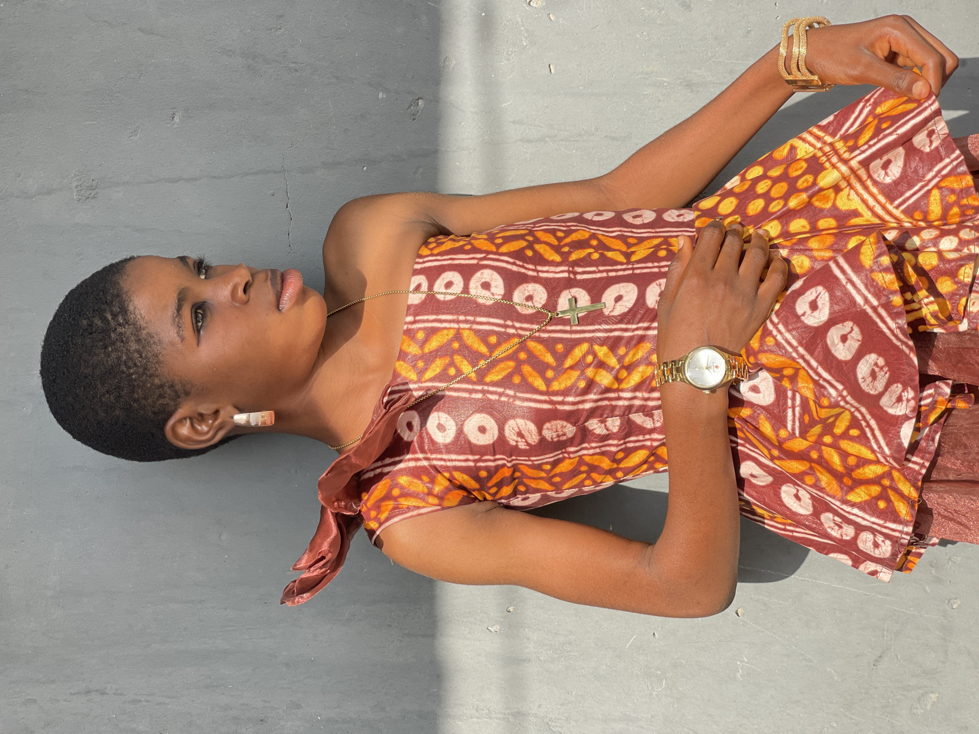 A young woman is depicted lying on a textured, grey concrete surface under bright, direct sunlight, casting strong shadows. Her body is positioned horizontally across the frame, while her head is turned to look upwards and slightly to her left. She has short, dark hair and dark skin, and her facial expression is calm and contemplative.
She is wearing a strapless or off-shoulder dress made from a fabric with a distinctive pattern in shades of reddish-brown, orange, and off-white. The pattern features a mix of circular motifs and abstract shapes, reminiscent of traditional West African textiles, which aligns with the Badagry, Nigeria context.
Her accessories include a delicate gold-colored chain necklace with a small cross pendant resting on her chest. On her left wrist, she wears a gold-colored watch with a light face, and on her right wrist, a large, ornate gold-colored cuff bracelet. A prominent, shiny silver-colored ear cuff or large earring is visible on her left ear. Her hands are positioned on or near her dress, with her right hand gently holding a fold of the fabric.
The bright sunlight and sharp shadows indicate a clear, sunny day, likely midday or early afternoon. No visible text is clearly discernible on her attire or accessories. The overall scene suggests a posed portrait outdoors, highlighting the woman and her vibrant dress.