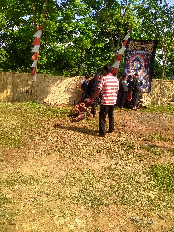The image depicts an outdoor scene in Plosorejo, Indonesia, under bright daylight. The foreground consists of an uneven, dry dirt ground with sparse green grass and minimal scattered debris. In the mid-ground, an individual wearing a red and white striped shirt and dark trousers stands with their back to the camera. To their left, another person is on the ground in a crouched or prone position, wearing dark, patterned attire and a head covering, appearing to be engaged in a performance or demonstration. Behind these individuals, a group of approximately 8-10 people, mostly in dark clothing, are gathered near a woven bamboo fence. A large dark blue banner displaying "PSHW-TM SUB RANTING" and a central circular emblem is prominently hung on the right side, attached to a vertical support pole. Two vertical poles adorned with red and white triangular pennant flags are visible, one on the left of the fence and another partially visible on the right. The bamboo fence, which spans horizontally across the background, has faint markings or graffiti on its left section, appearing to read "SENA". Numerous trees with dense green foliage are visible behind the fence and banner, under a clear sky. Shadows are cast to the left, indicating the sun's position.
