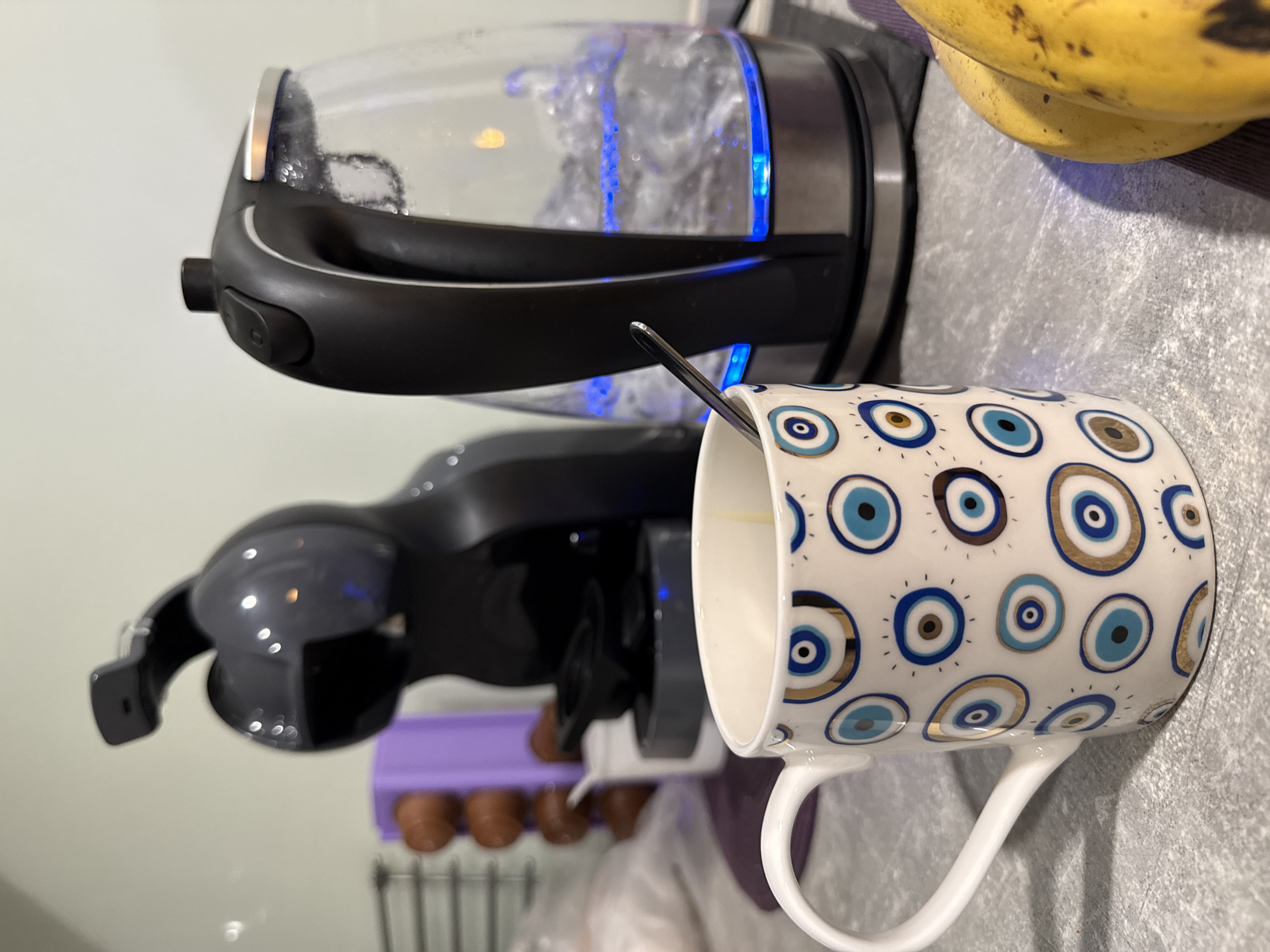 A close-up shot depicts a kitchen countertop scene. A clear glass electric kettle with black accents is actively boiling, with visible blue LED illumination indicating the heating process. A white mug, adorned with a repeating blue and gold evil eye pattern, sits adjacent to the kettle, containing a small amount of light-colored liquid and a stainless steel spoon. A dark grey Nespresso machine is also present in the background. Further back, partly obscured, is a purple dish rack holding what appear to be sausages. The background includes hints of white walls and what seems to be a portion of bananas are also on the top right side of the picture. There's no indication of Sofia, Bulgaria or people visible in the frame.