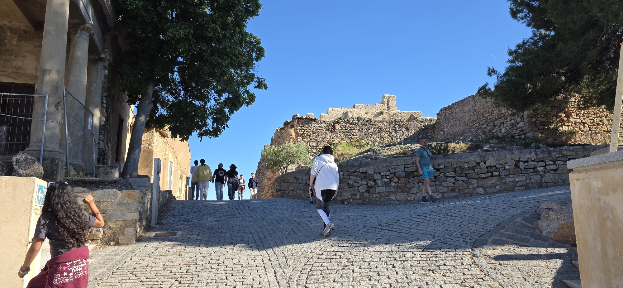 The image depicts a cobblestone street leading uphill towards a stone structure, likely ruins, under a clear blue sky in Sagunto, Spain. Several people are walking up the street, with one person facing away from the camera in the foreground, and a group of people further up the street. There is also an individual standing near the stone wall on the right. Buildings and trees flank the street on both sides. The light and shadows suggest it is daytime. A partially obstructed building with columns is visible on the left.