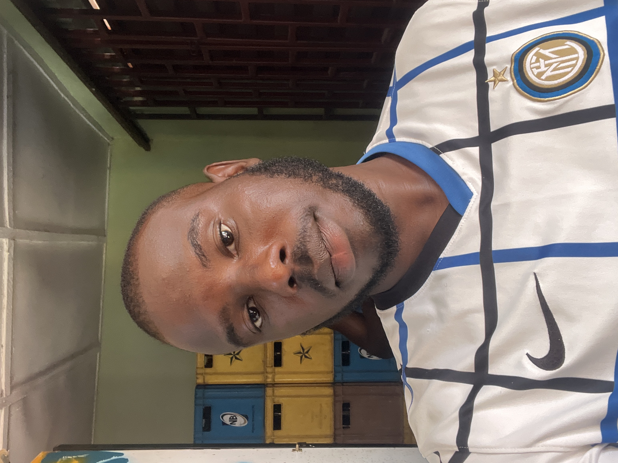 A medium shot features a dark-skinned man with a shaved head and full beard, wearing a white soccer jersey with black and blue grid lines and a Nike swoosh logo. The jersey also has a circular emblem with a gold star above it. He is looking directly at the camera. Behind him, there is a stack of plastic crates in blue, yellow, and brown. A wall is visible on the left and green wall behind crates. The roof seems to have brown wood. Given the context, it is presumed that the location is Jos, Nigeria.