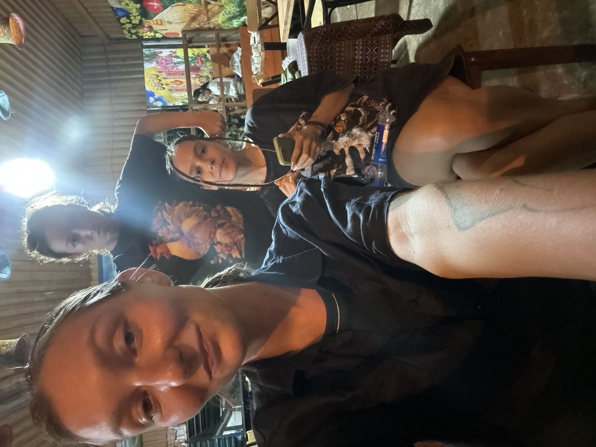 This image is a selfie taken indoors at what appears to be a cafe or communal area in Mù Cang Chải, Vietnam, likely in the evening or at night given the artificial lighting.
Three individuals, appearing to be young adults or women, are the main subjects. The person in the foreground is taking the selfie, showing her face with a slight smile and a portion of her arm, where a tattoo is visible. Behind her and to the left, another person with curly dark hair is partially visible, her face obscured by glare. The third individual is further back on the right, seated and holding a smartphone. She has long dark hair and is wearing a dark t-shirt with a graphic design. Next to her is an "AQUAFINA" water bottle. Another person is dimly visible sitting at a table in the background.
The setting is characterized by a rustic charm, with corrugated metal walls or ceiling. The background features colorful wall hangings, possibly tapestries or paintings, depicting floral and foliage motifs, as well as woven basket decorations, indicative of traditional Vietnamese craftsmanship. The overall atmosphere appears relaxed and social.