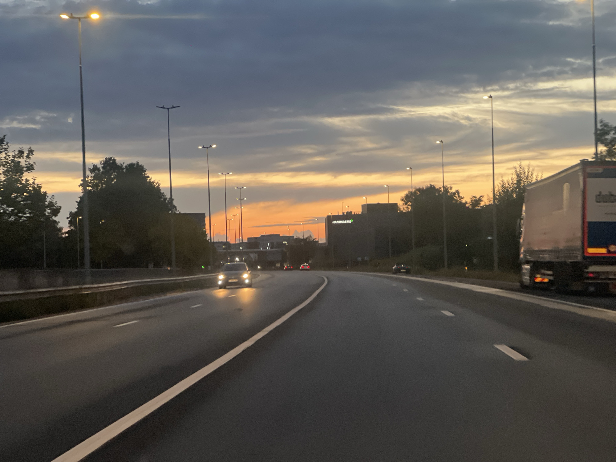A low-angle, medium shot captures a highway scene at dusk in Unknown, Unknown. The sky displays a dramatic sunset with dark, cloudy formations above bands of orange and yellow light near the horizon. A line of streetlights stretches along the left side of the road, some illuminated. Trees line the median and the right side of the highway.
On the road, several vehicles are visible. In the foreground on the left lane, a car with its headlights on is approaching. Further ahead, another car is visible in the right lane. A large truck is traveling in the right lane on the far right side of the image. A building with "HANDSAEM B" visible on its facade is located in the background. The asphalt road is wet and reflects the surrounding lights.