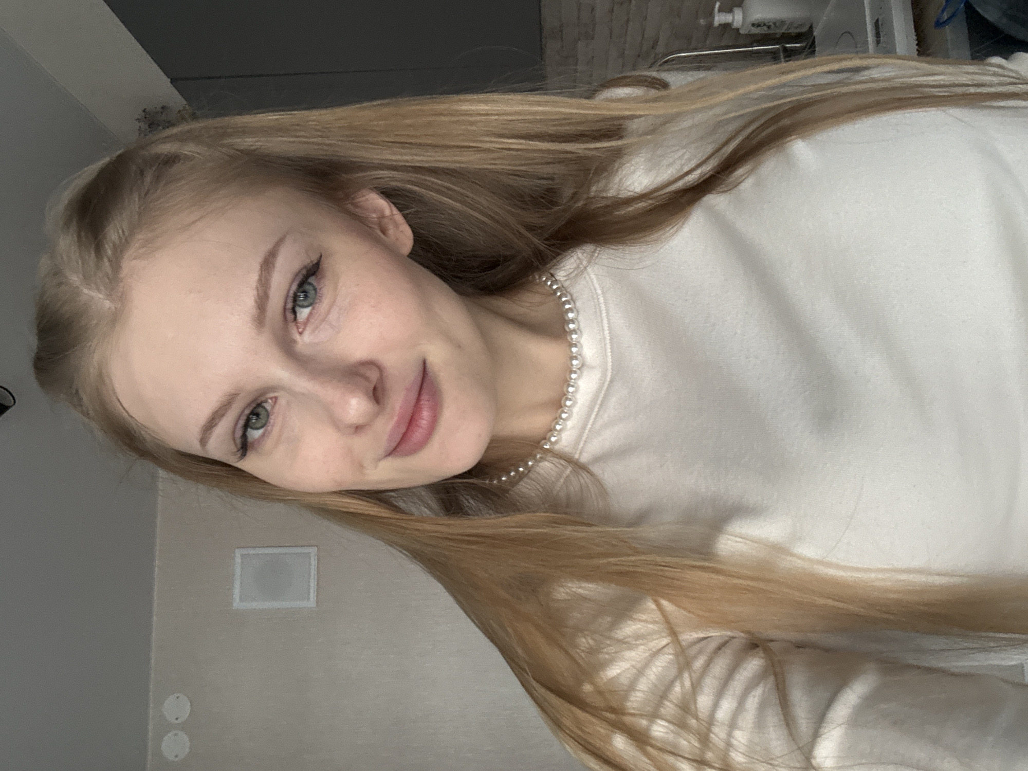 A young woman with long, blonde hair and green eyes is pictured in a close-up, selfie-style shot. She is wearing a light-colored, long-sleeved shirt and a pearl necklace. Her lips are slightly parted, and she is smiling gently. The background appears to be an indoor setting, possibly an apartment, with a light-colored wall, a framed white object on the wall, and two circular white fixtures on the ceiling. A gray cabinet or wall is visible on the right side of the image, along with what looks like a countertop with a bottle of soap or lotion. The scene is illuminated by soft, natural light, suggesting it was taken during the day. There is no visible indication of the city of Perm, Russia within the image itself.