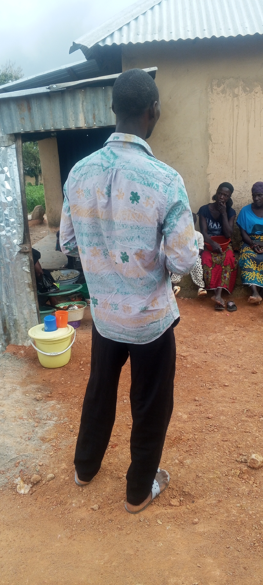 In an outdoor scene on a dirt ground, a man stands with his back to the camera, facing away from the viewer. He is wearing a light-colored, short-sleeved button-up shirt with a pattern of green clover-like shapes and stripes in shades of blue and peach. He also wears dark pants and light-colored sandals. In the background, to the right, three women are seated on the ground in front of a tan-colored building with a corrugated metal roof. The woman on the left wears a dark shirt and a red and blue patterned skirt. The middle woman has on a dark blue shirt and a red and yellow patterned skirt. The woman on the right is in a bright blue top and a yellow and blue patterned skirt. A red bowl sits on the lap of the middle woman. To the left of the man, near an opening in a corrugated metal structure, a yellow bucket with a white rim holds a blue cup and an orange cup. Several other containers and bowls are visible behind the bucket. The environment appears to be rural or in a developing area, with loose dirt and scattered rocks on the ground, and vegetation visible behind the structures. The sky is overcast.