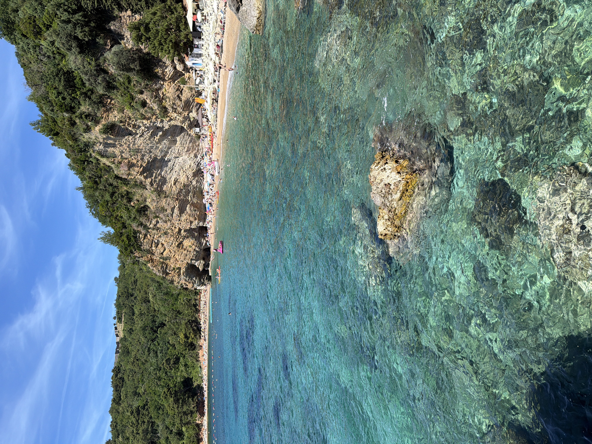 This is a scenic view of a beach in Budva, Montenegro, likely Mogren Beach due to the presence of a cliff face with visible rock formations and dense vegetation. The water is remarkably clear and a vibrant turquoise, allowing visibility of the rocky seabed and scattered boulders. The foreground features a large, weathered rock protruding from the water, partially submerged.
The beach itself is populated with numerous sunbathers, appearing as small figures scattered across the sand. There are rows of white lounge chairs and umbrellas set up on the beach, suggesting a well-organized tourist area. A few people are in the water, some swimming, and others on inflatable floats, including a bright pink one. Kayaks and paddleboards are also visible near the shore.
The background is dominated by a steep, layered cliff face covered in lush green trees and vegetation. At the top of the cliff, a few structures are faintly visible, possibly part of an old fortress or ruins. The sky is a clear, bright blue with wispy white clouds. The overall atmosphere is one of a sunny, active beach day in a picturesque coastal location.
