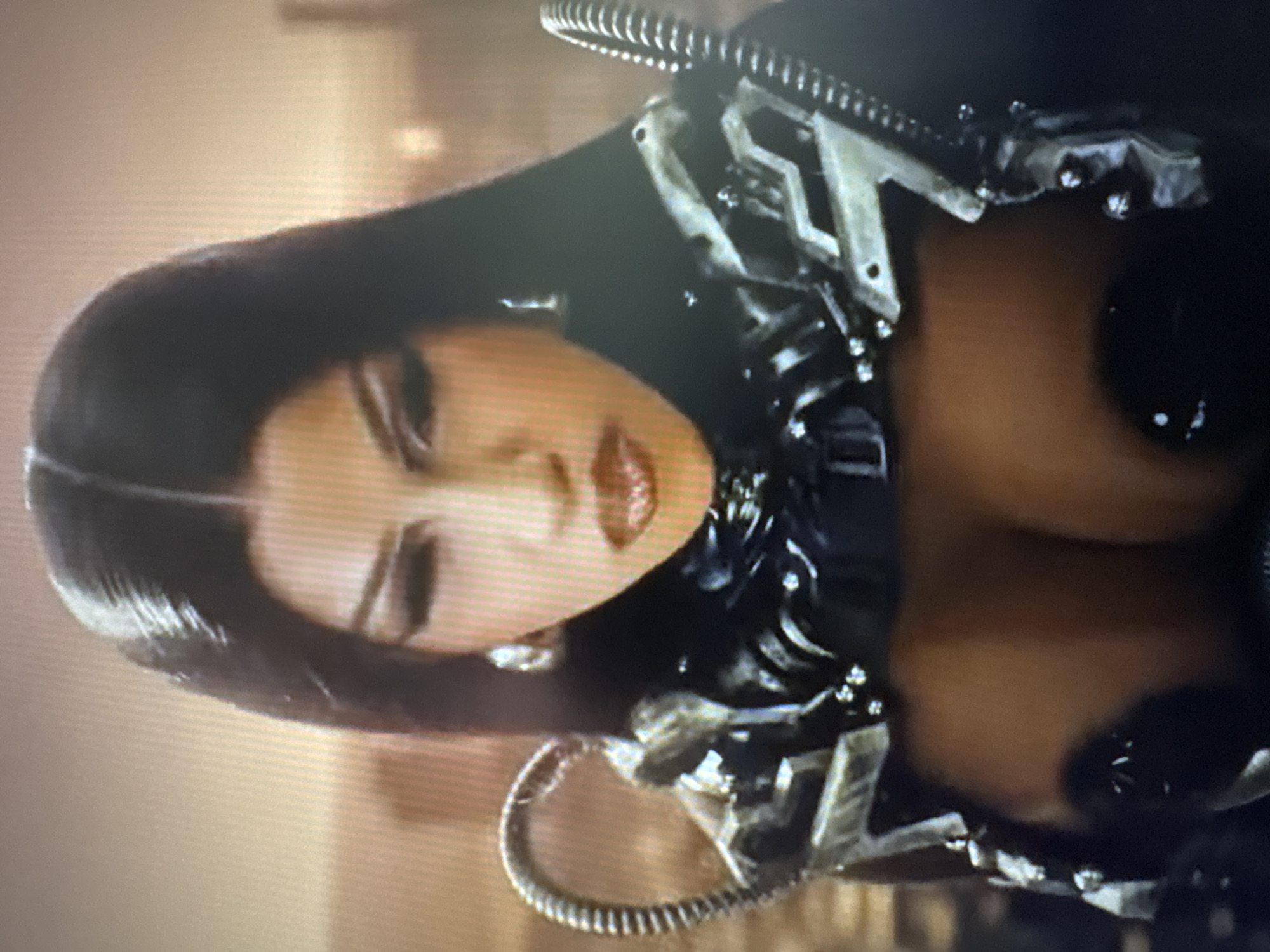 The image is a close-up, medium shot of a woman. She has long, straight, dark hair parted in the middle. Her makeup includes prominent eyeliner and shimmery lip gloss. She is wearing large hoop earrings and a futuristic-style costume with metallic shoulder pieces featuring ribbed tubes. The background appears to be an urban landscape, possibly a city at dusk, with blurred buildings and a muted color palette. There is no identifiable information suggesting the image is located in Sofia, Bulgaria.