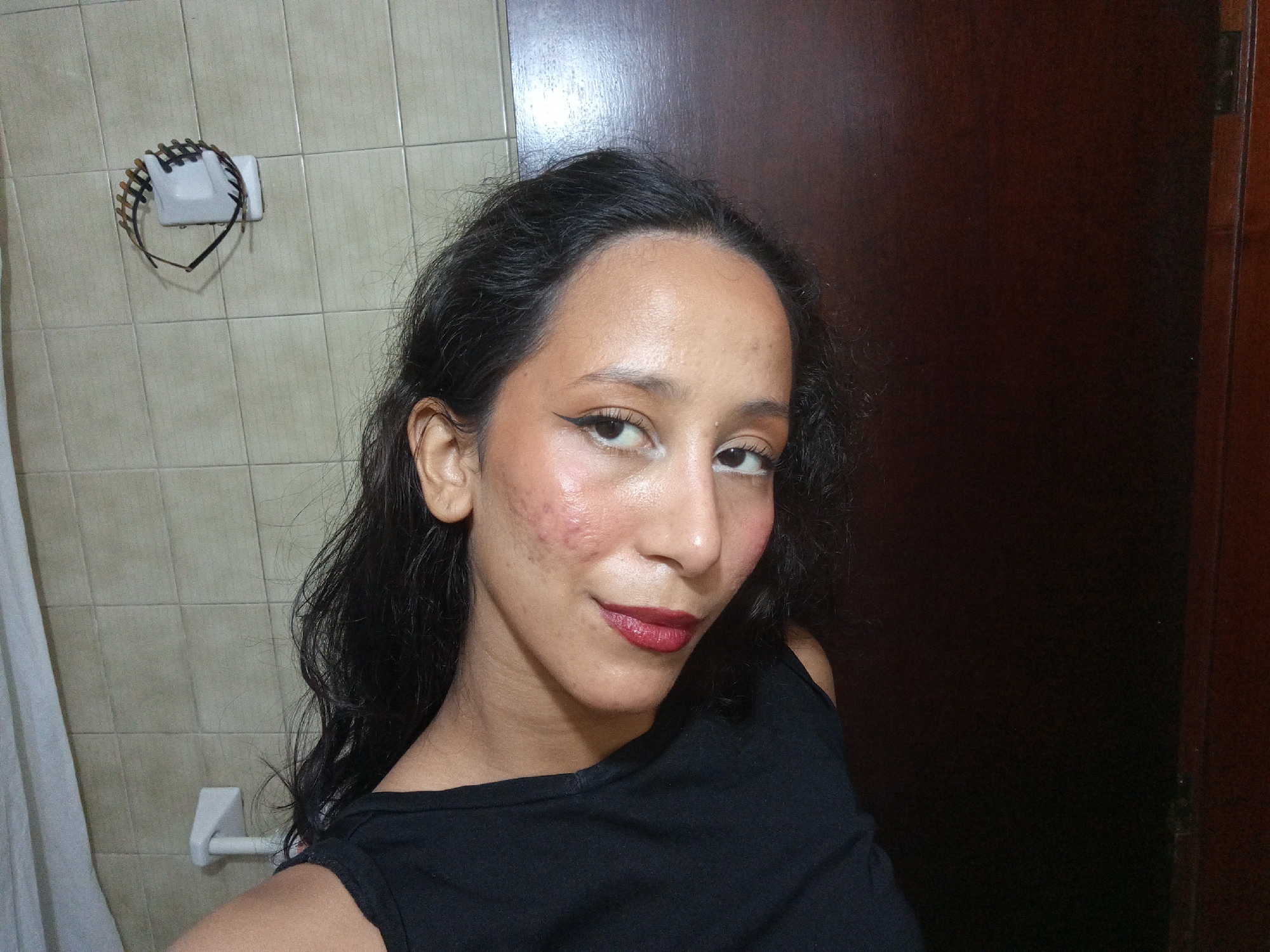 A woman is standing in a bathroom. She has long, dark, wavy hair and is wearing a dark top. She is facing the camera with her head tilted slightly to her right. She is wearing makeup, including eyeliner and lipstick. The background includes a tiled wall with a headband and a towel rack. A dark wooden door is visible in the background to the right. It is not possible to verify the location of Porlamar, Venezuela.