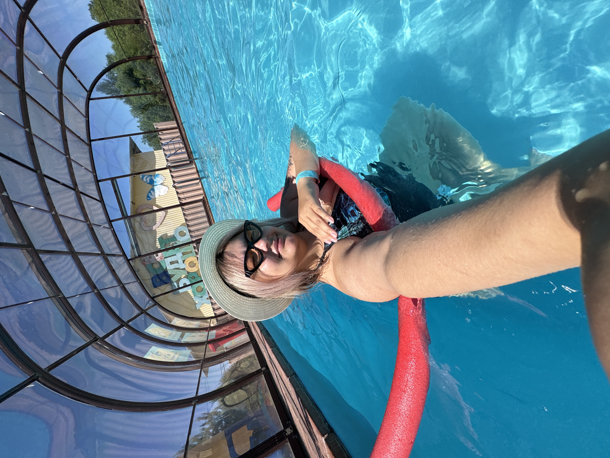 A woman wearing a straw hat and sunglasses floats in a swimming pool under a clear glass enclosure. She is holding a red pool noodle. The pool has bright blue water with visible ripples. In the background, there is a building with colorful signage and trees outside the enclosure. The woman is taking a selfie.