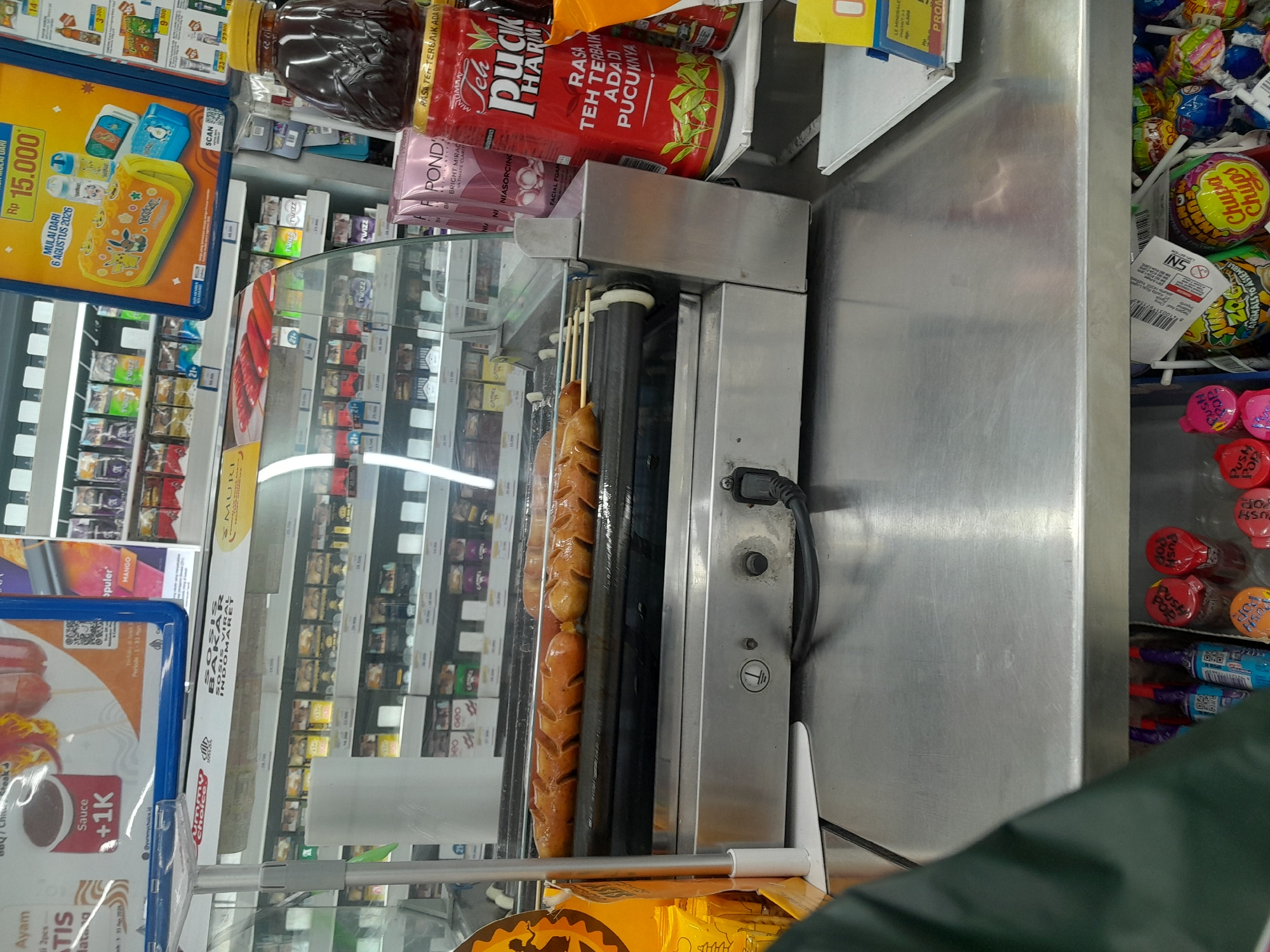 The image depicts an interior view of a convenience store, likely a minimarket, with a focus on a hot dog roller grill located in the foreground. The grill is loaded with several rows of cooked sausages, some appearing to be hot dogs or similar processed meat products, arranged on a rotating spit. The grill itself is made of stainless steel and has a transparent glass cover. To the right of the grill, on a shelf, are several products including a large bottle of iced tea with Indonesian text and packaging, and some smaller pink packages of what appears to be facial cleanser. In the background, refrigerated display cases are visible, stocked with various food and beverage items, including cigarettes, snacks, and drinks. A promotional sign with an image of cartoon characters and pricing information in Indonesian is also visible in the upper right portion of the frame. The overall environment suggests a commercial retail setting where prepared foods are sold alongside packaged goods. The location is specified as Desa Ponokawan, Indonesia, indicating a rural or semi-urban area within Indonesia. The image does not contain any visible people.