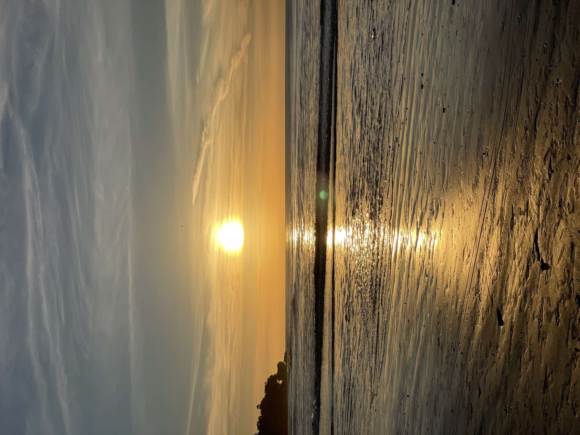 The image captures a sunset scene at a beach. The sun is positioned near the horizon, casting a golden glow across the sky and reflecting on the wet sand. The sky displays a gradient from light blue to orange-yellow. The sea is visible as a strip, with gentle waves. The foreground shows the sandy beach, with patterns and textures created by the receding tide. There are some dark patches of vegetation near the horizon. The location is in the city of Unknown, Unknown.