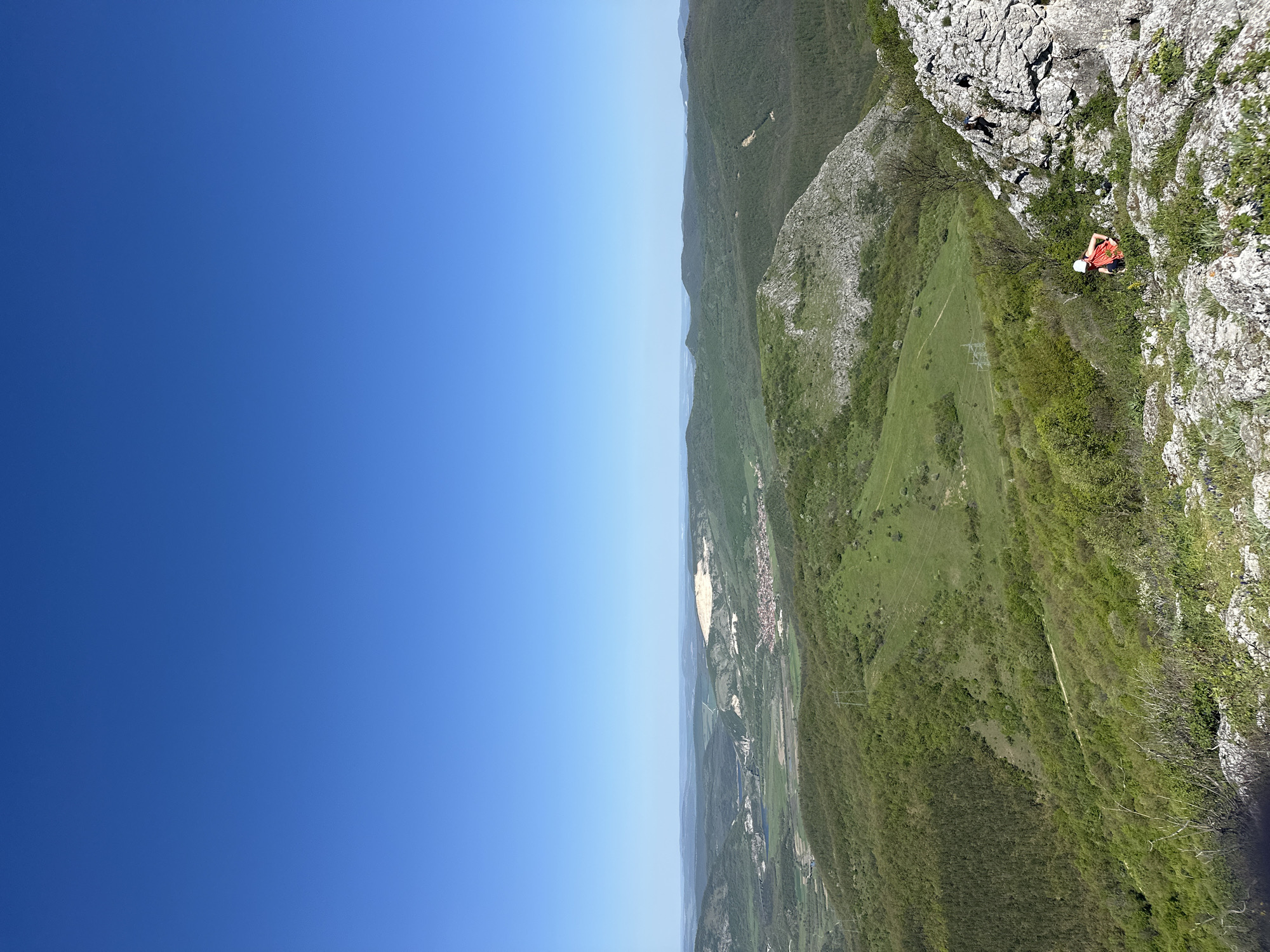 The image depicts a climber resting on a rock face in a mountainous area. The location is possibly near Bilka, Bulgaria, judging by the landscape. A person wearing a white helmet and an orange shirt is resting on a rock formation. The background shows a valley with fields, vegetation, and a village. The sky is clear and blue. The image is taken from a high angle, showing the vastness of the landscape.
