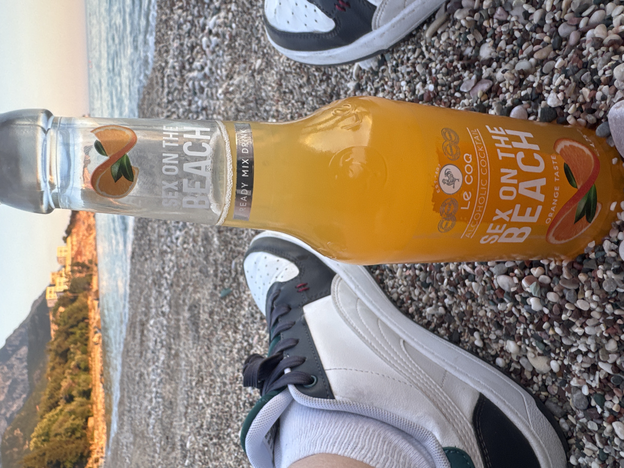 The image shows a bottle of "Sex on the Beach" cocktail placed on a pebble beach. Two pairs of sneakers are visible, presumably worn by people enjoying the beach. The background features the sea and a coastal landscape with buildings and trees. The location is likely Kamenovo, Montenegro, based on the coastal architecture and pebble beach environment.