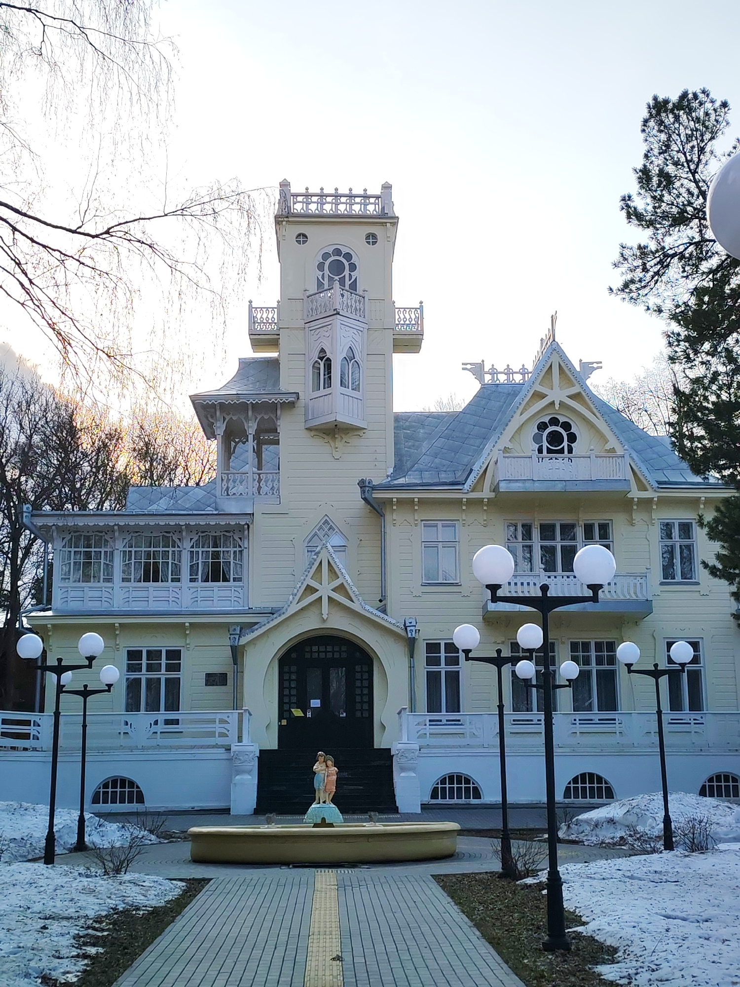 The image shows the Dacha of Bashenin, an architectural landmark located in Sarapul, Russia. The building is a two-story light-yellow structure with a prominent central tower and intricate white trim details, including balconies, decorative eaves, and window designs. A paved walkway leads to a fountain with a small statue in front of the building's entrance. Several globe-shaped streetlights line the path and the edges of the grounds. Patches of snow are visible on the ground, suggesting a late winter or early spring setting. Bare trees are visible in the background on the left, and evergreen trees are on the right. The sky is overcast.