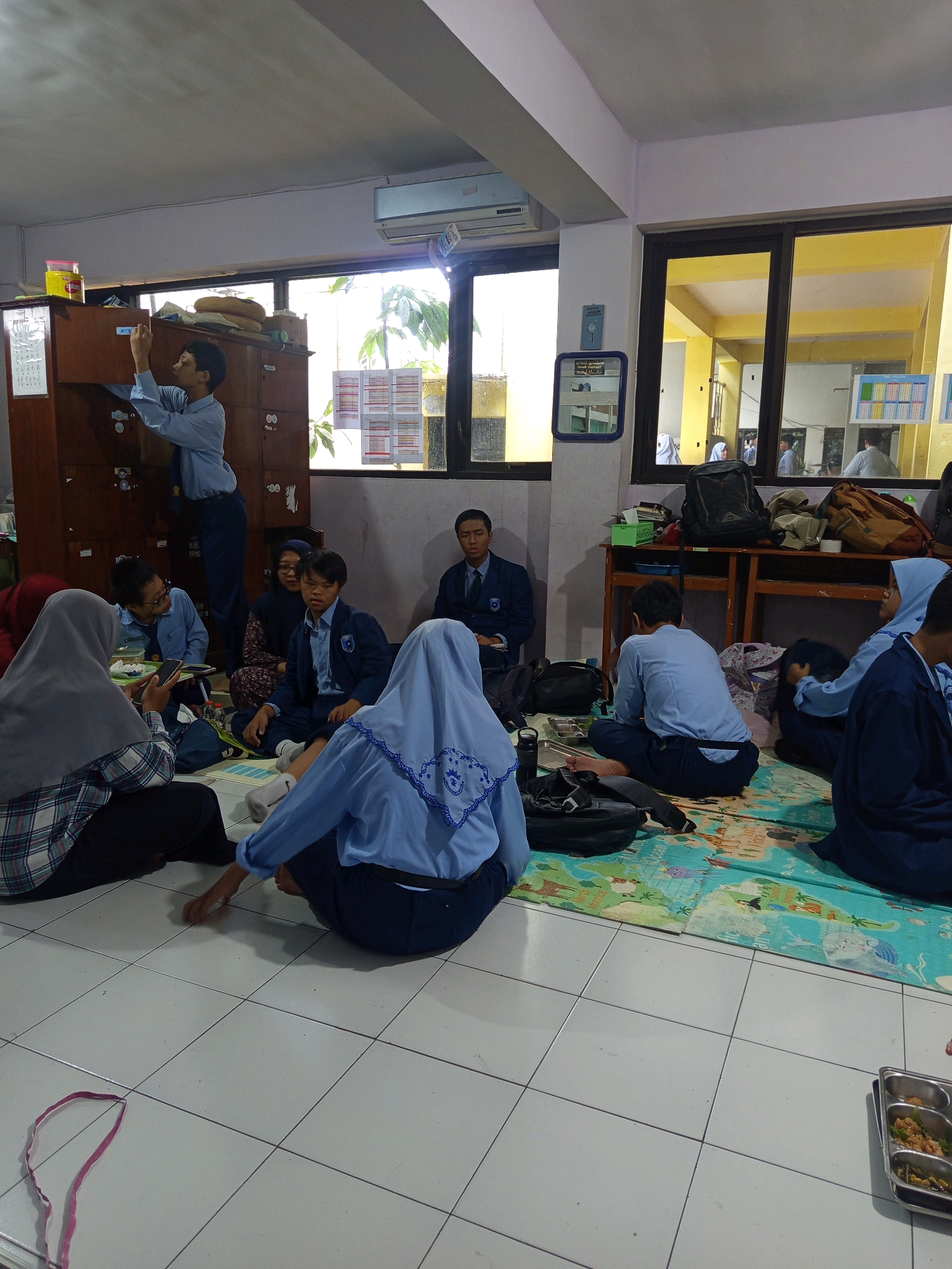 The image depicts a group of students, some in school uniforms, gathered in a classroom or common area, possibly in Malang, Indonesia. The students are seated on the floor, some facing a wall with lockers, while others are engaged in conversation or looking at their phones. One student is standing and appears to be reaching into a locker. The room has large windows with views of an outdoor area with greenery. There are tables and chairs in the background, and various bags and personal belongings are scattered around the floor. The overall atmosphere suggests a casual gathering or study session.