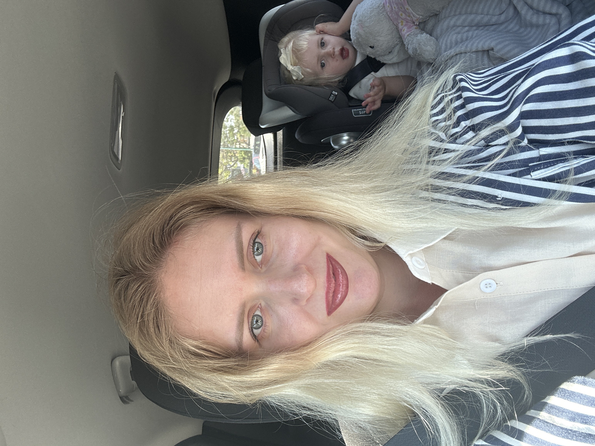 A woman with blond hair and blue eyes is taking a selfie in the passenger seat of a car. She is wearing a beige shirt and a blue and white striped jacket. Her lips are red. A baby is in a car seat in the back. The baby has blond hair and is holding a stuffed animal under a blanket. The car is in Petah Tikva, Israel. Trees are visible outside the window.