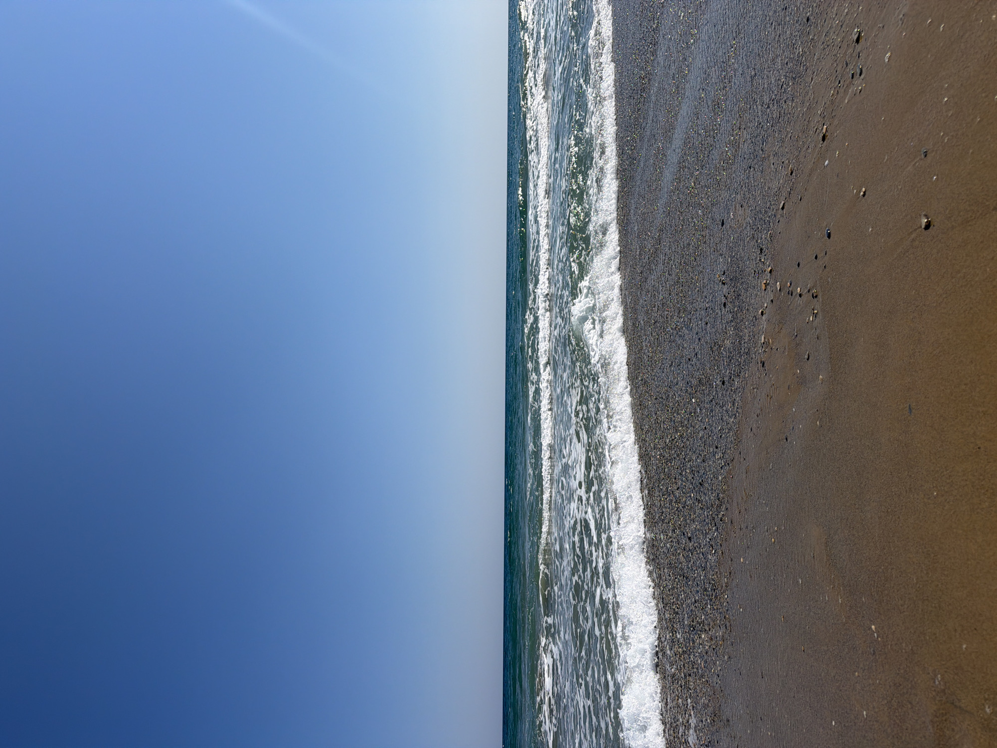 The image depicts a serene beach scene in Georgioupoli, Greece, under a clear, bright blue sky. The foreground is dominated by the wet sand of the beach, with small pebbles scattered across its surface. A gentle wave with white foam is seen receding from the shore, leaving the pebbled area glistening. Beyond the shore, the turquoise waters of the sea stretch out to the horizon, with a few more waves breaking further out. The horizon line is sharp and defined, separating the sea from the vast expanse of the sky. Sunlight glints off the surface of the water, creating a sparkling effect. No people or man-made structures are visible in the frame, emphasizing the natural beauty of the location. The overall impression is one of tranquility and a perfect summer day at the seaside.