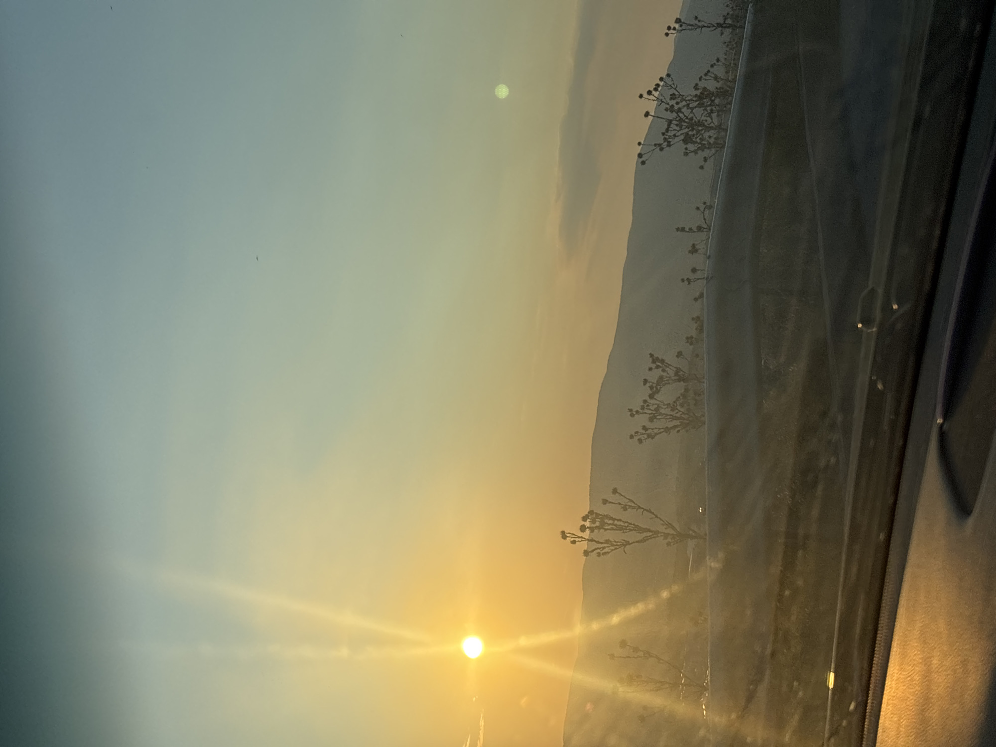 A low-angle, medium shot captures a sunset over a mountainous landscape, viewed from inside a vehicle. The sun, a bright orange orb, is positioned in the upper left quadrant of the image, casting long rays of light that streak across the sky. The sky transitions from a warm yellow near the horizon to a pale blue at the top. Silhouetted against the sunset are rolling hills and scattered dry, thorny plants in the middle ground. The foreground consists of a car's dashboard and windshield, with reflections of the sunset visible on the glass. The overall scene conveys a sense of calm and natural beauty during a sunset.