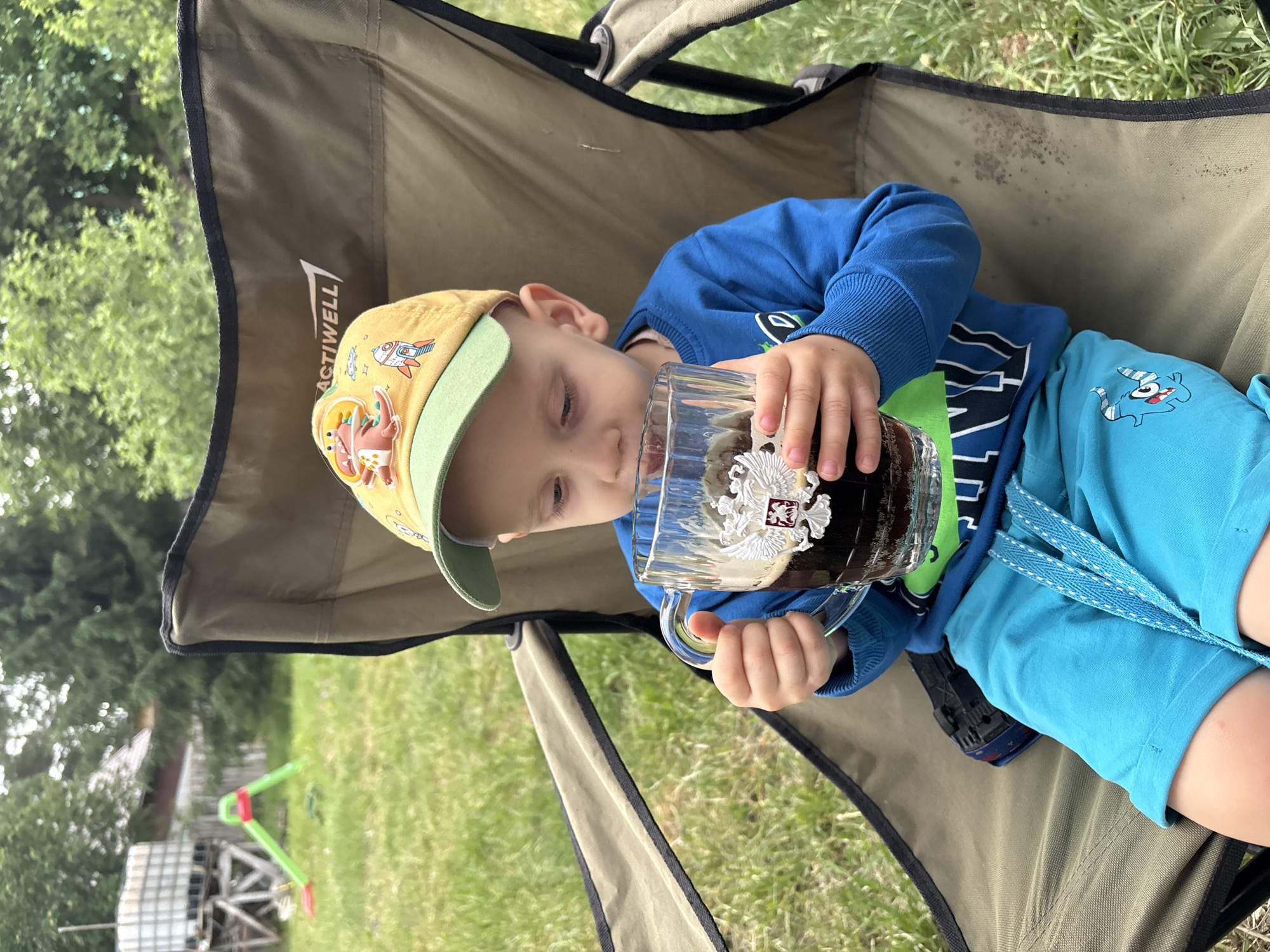 A young child wearing a yellow hat, blue long-sleeved shirt, and blue shorts is sitting in a camping chair and drinking from a large glass mug. The mug appears to contain a dark liquid. The child is holding the mug with both hands. The camping chair is green and the background is a grassy area with trees. There's a seesaw and a large tank-like object in the background. The location is Chaikovsky, Russia.