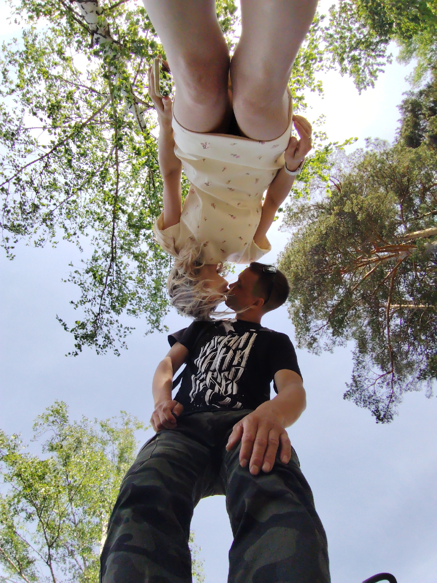 A low-angle photograph shows a couple kissing, with the woman upside down. She is wearing a light-colored dress with a floral pattern and a white watch, holding a smartphone in her right hand. The man is wearing a black t-shirt with a white graphic and camouflage pants. They are positioned under trees, with the sky visible in the background. The photo was taken in the city of Chaikovsky, Russia.