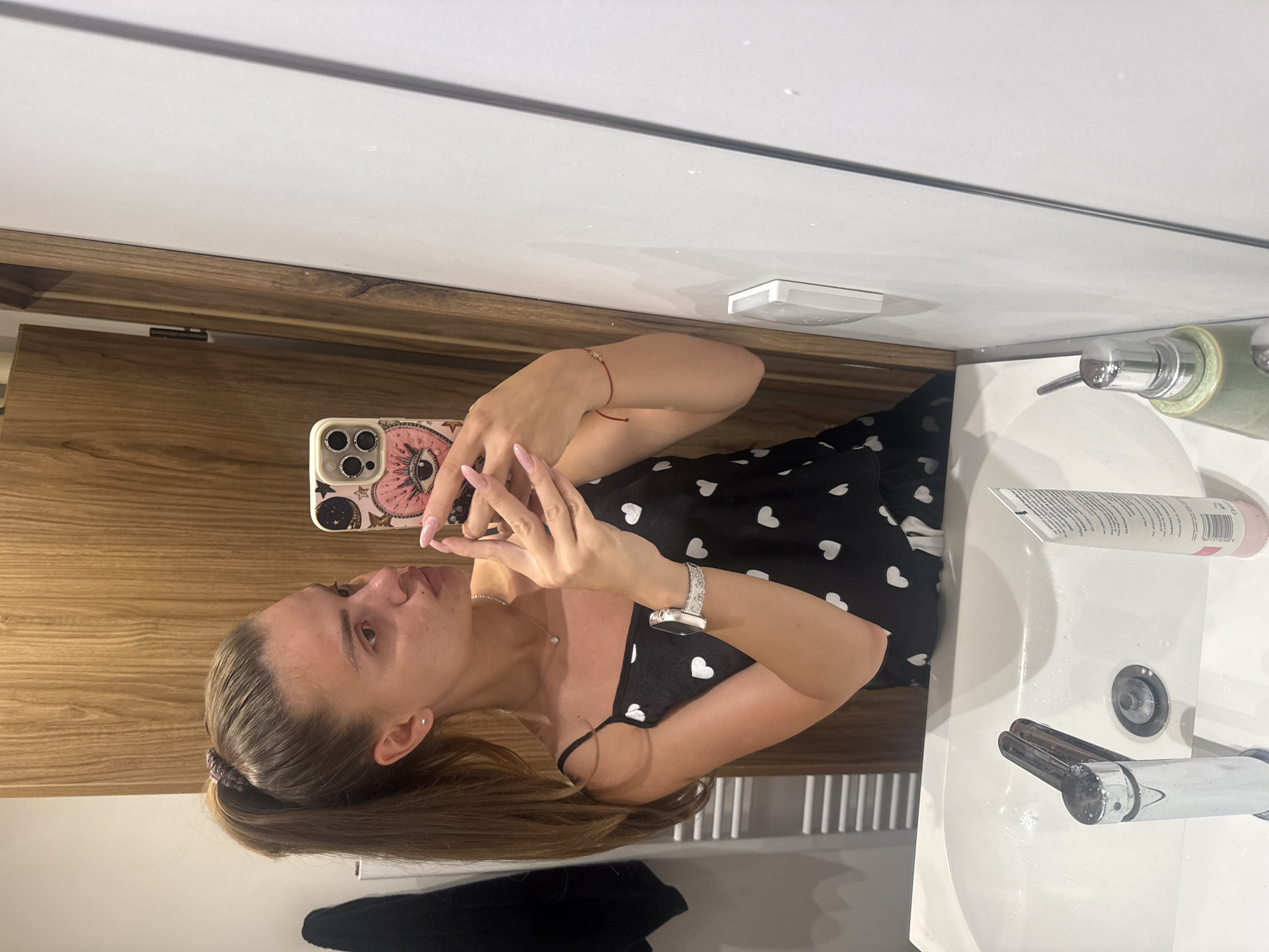 The image shows a young woman with her hair in a high ponytail, taking a selfie in a bathroom mirror. She is wearing a black tank top with a white heart pattern. On her left wrist, she has a silver smartwatch, and on her right wrist, a thin red string bracelet. Her nails are painted a light pink. She is holding a smartphone with a pink case decorated with astrological symbols and an eye motif.
The bathroom appears to have a white sink with a chrome faucet and drain. To the right of the sink, there is a white tube, likely a cosmetic product, and a green dispenser bottle. The background shows a wooden cabinet and a white tiled wall. The overall lighting suggests an indoor setting, possibly a residential bathroom.
The image was taken in the city of Plovdiv, Bulgaria.
