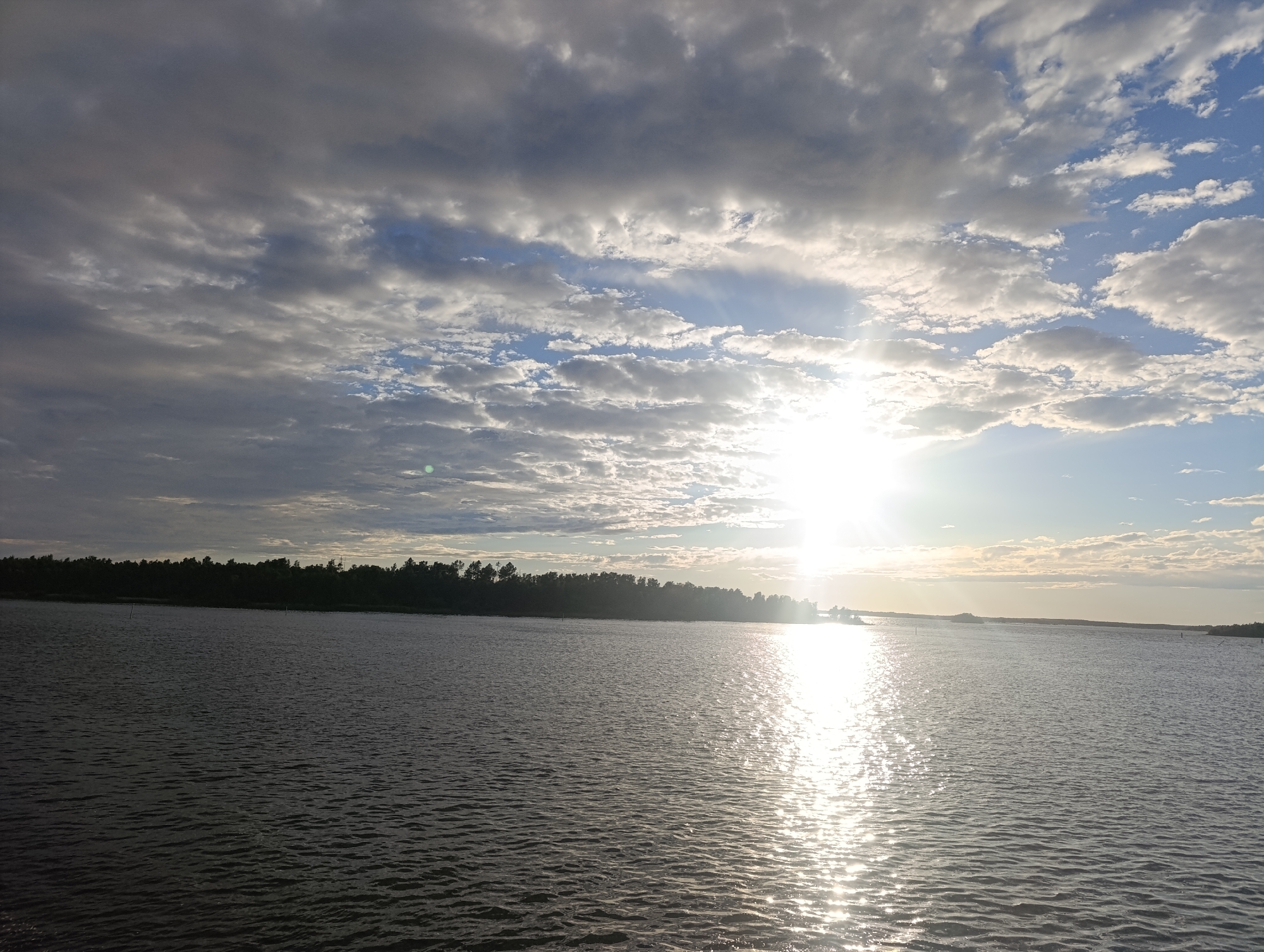 The image depicts a wide body of water under a cloudy sky, with the sun shining brightly and reflecting off the water's surface. A treeline forms a dark silhouette along the horizon. The scene appears to be a natural landscape rather than an urban environment, making it unlikely to be located within the city of Jakobstad, Finland, which is known for its maritime heritage and industrial areas. The overall impression is one of a serene, natural setting, possibly a lake, river, or coastal area at dusk or dawn.