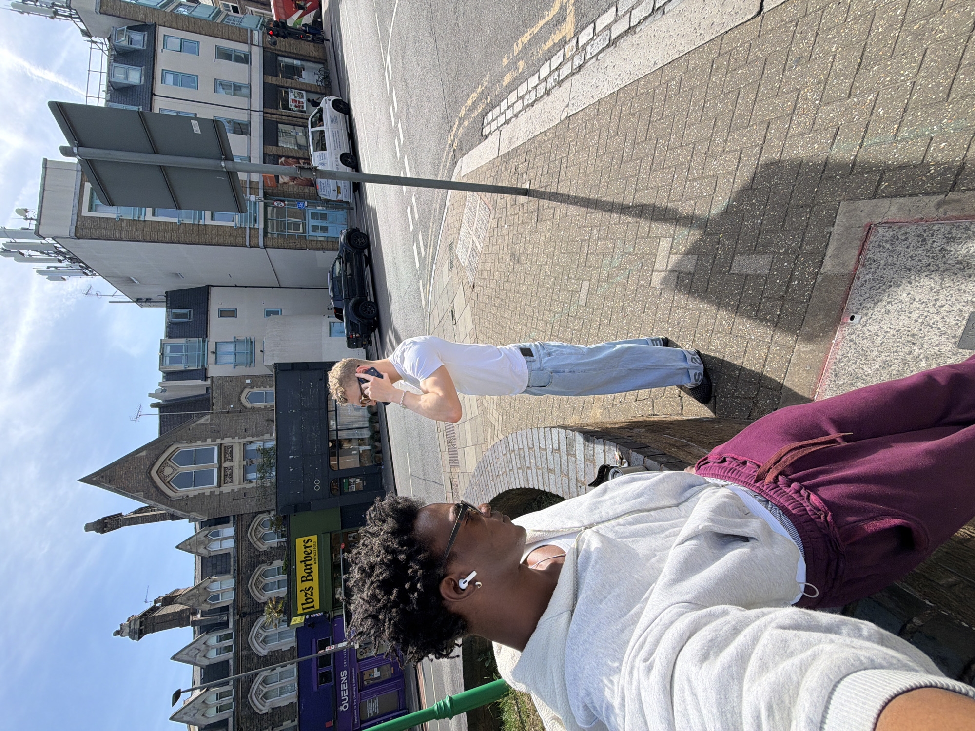 The image shows two individuals outdoors, likely in London, United Kingdom. A person with dark hair and sunglasses is wearing a light-colored hoodie and maroon sweatpants, with white earbuds in their ear. A second person, with blonde hair and sunglasses, is wearing a white t-shirt and light blue jeans, holding a phone to their ear. They are standing near a street, with buildings in the background, including a building with the sign "Ibz's Barbers." Cars are parked on the street.