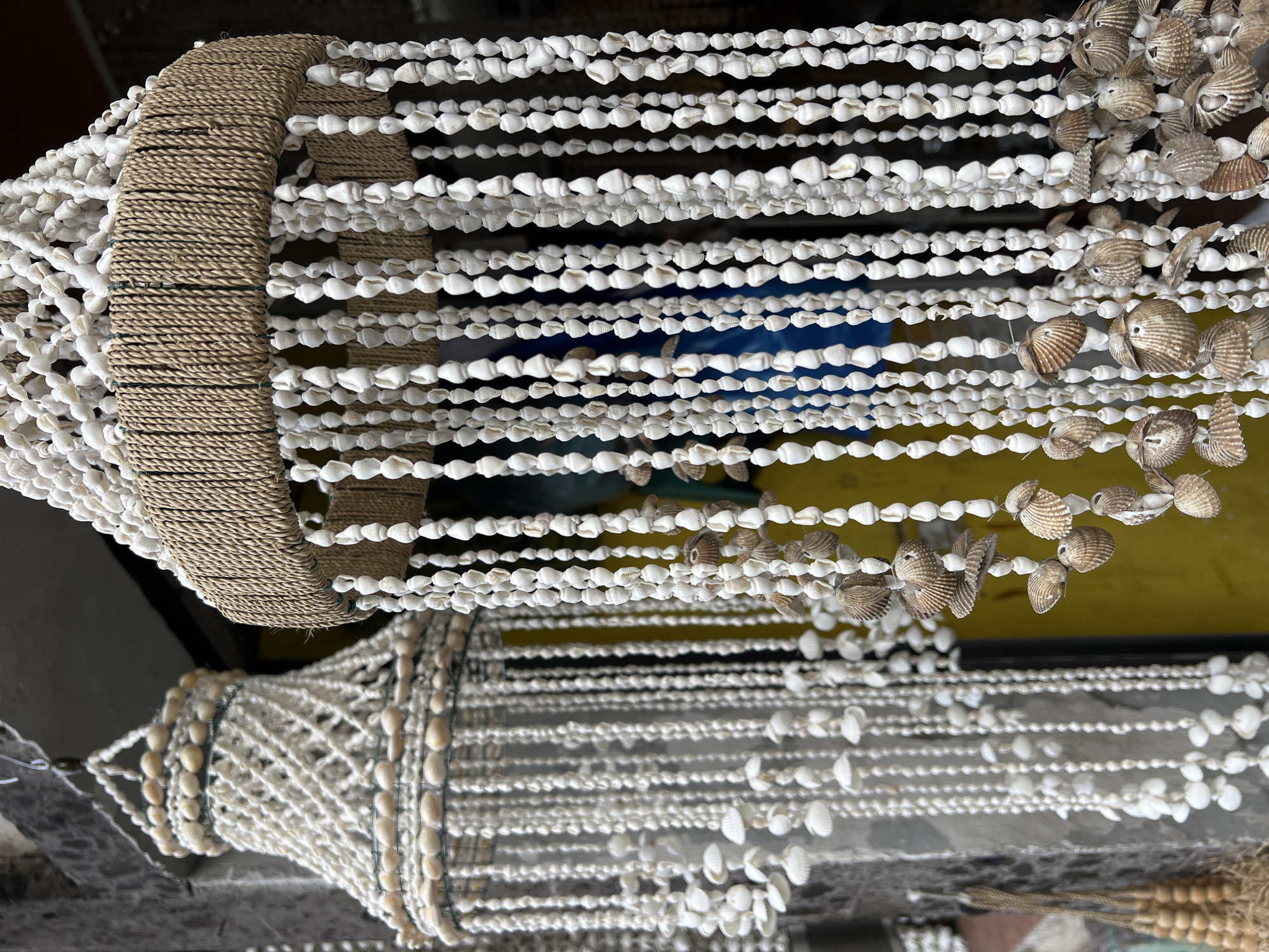 The image captures two shell chandeliers, one hanging slightly higher than the other. The upper chandelier features a woven, rope-like circular frame in a natural brown color. Strands of white shells dangle from this frame, creating a curtain-like effect. Some strands also incorporate brown shells. The chandelier below is structured differently; it has a more geometric, cage-like construction made of wire, also adorned with white shells. The background reveals a blur of colors, with yellow and blue tones suggesting an interior space. The picture was taken in the Ubud District of Indonesia.