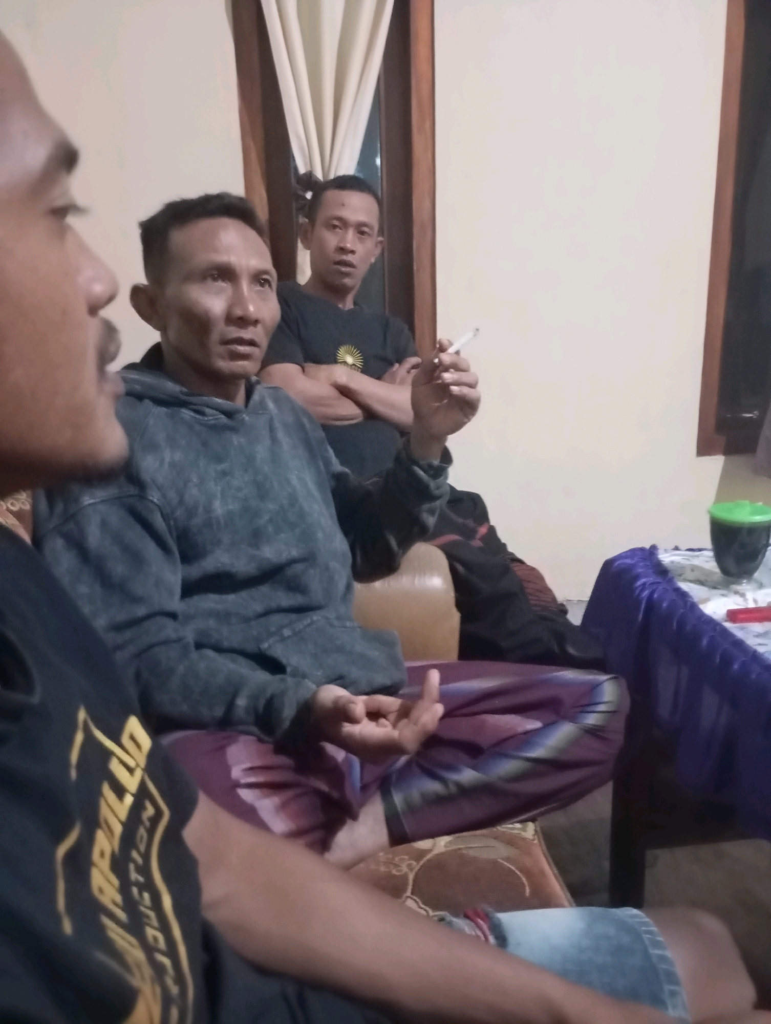 Three adult males are seated indoors, possibly in a living room, with a neutral-colored wall and a wooden-framed window behind them. The man in the center is wearing a dark gray hooded sweatshirt and appears to be speaking. To his right, another man in a black t-shirt with a yellow graphic is holding a lit cigarette and looking towards the left. In the foreground, a third man, mostly out of focus, is wearing a black t-shirt with a yellow graphic and is facing to the right. The room contains a brown couch and a purple draped surface. The overall lighting suggests an indoor setting, likely in the evening. The image does not provide specific clues to pinpoint the location within Wonogondo, Indonesia.