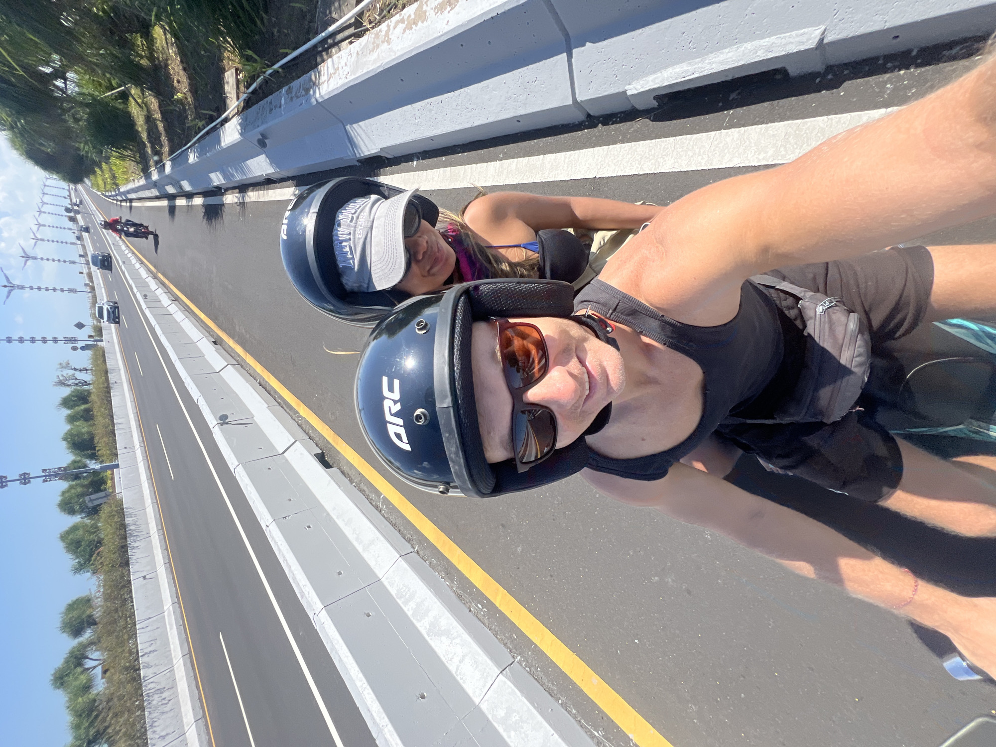 Two people are riding a motorcycle on a divided highway under a clear blue sky. The person in the foreground, wearing a black tank top and black helmet with "ARC" in white lettering, is taking a selfie. They are wearing sunglasses and a black fanny pack around their waist. Behind them, a person wearing a blue bikini top, a pink scarf, a grey baseball cap with "NEW YORK" in blue lettering, and a black helmet is visible. They are both looking towards the camera. The highway has a yellow line on the left and white lines dividing the lanes. Concrete barriers separate the lanes and the shoulder. Lush green vegetation and trees line the right side of the highway, and in the distance, several tall, decorative lampposts or sculptures are visible. Other vehicles, including cars and motorcycles, are also present on the highway in the background. The location appears to be on the Mandara Toll Road in Kuta Selatan, Indonesia, given the highway infrastructure and surrounding tropical vegetation.
