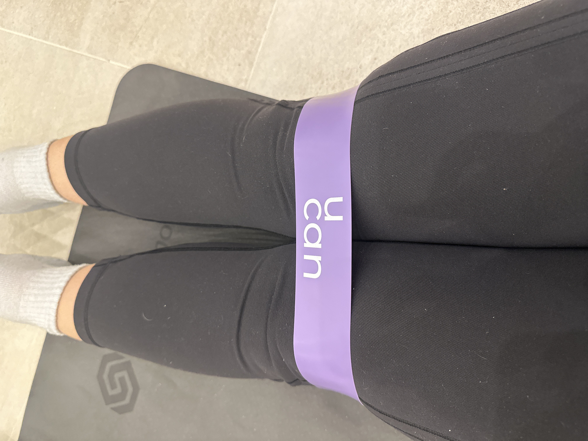 A close-up shot shows a person's legs resting on a gray yoga mat placed on a tile floor. The person is wearing black leggings and white socks. A light purple resistance band with the text "U Can" printed on it is placed around the thighs, just above the knees. The text on the band is oriented vertically. The location of the scene is not specified.