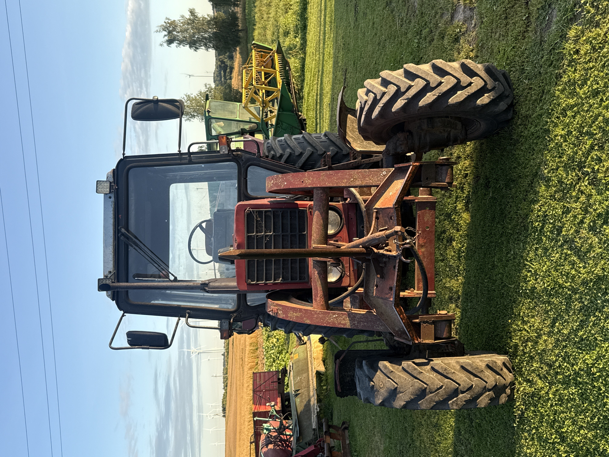 This is a front-facing, medium shot of a red tractor parked on a grassy field at sunset. The tractor is the primary subject, with its large, deeply treaded tires prominent in the foreground. The tractor has a metal cab with large glass windows and a rearview mirror visible on the driver's side. A black exhaust pipe extends vertically from the top of the cab. Attached to the front of the tractor is a metal frame with a hitching mechanism. In the background, a yellow and green agricultural machine is parked, along with a red trailer on the left. Beyond the machines, a field of golden wheat stretches towards the horizon, where several wind turbines are visible against a blue sky with scattered clouds. The setting sun casts long shadows across the grassy field. The image appears to have been taken in Vikraičiai, Lithuania.