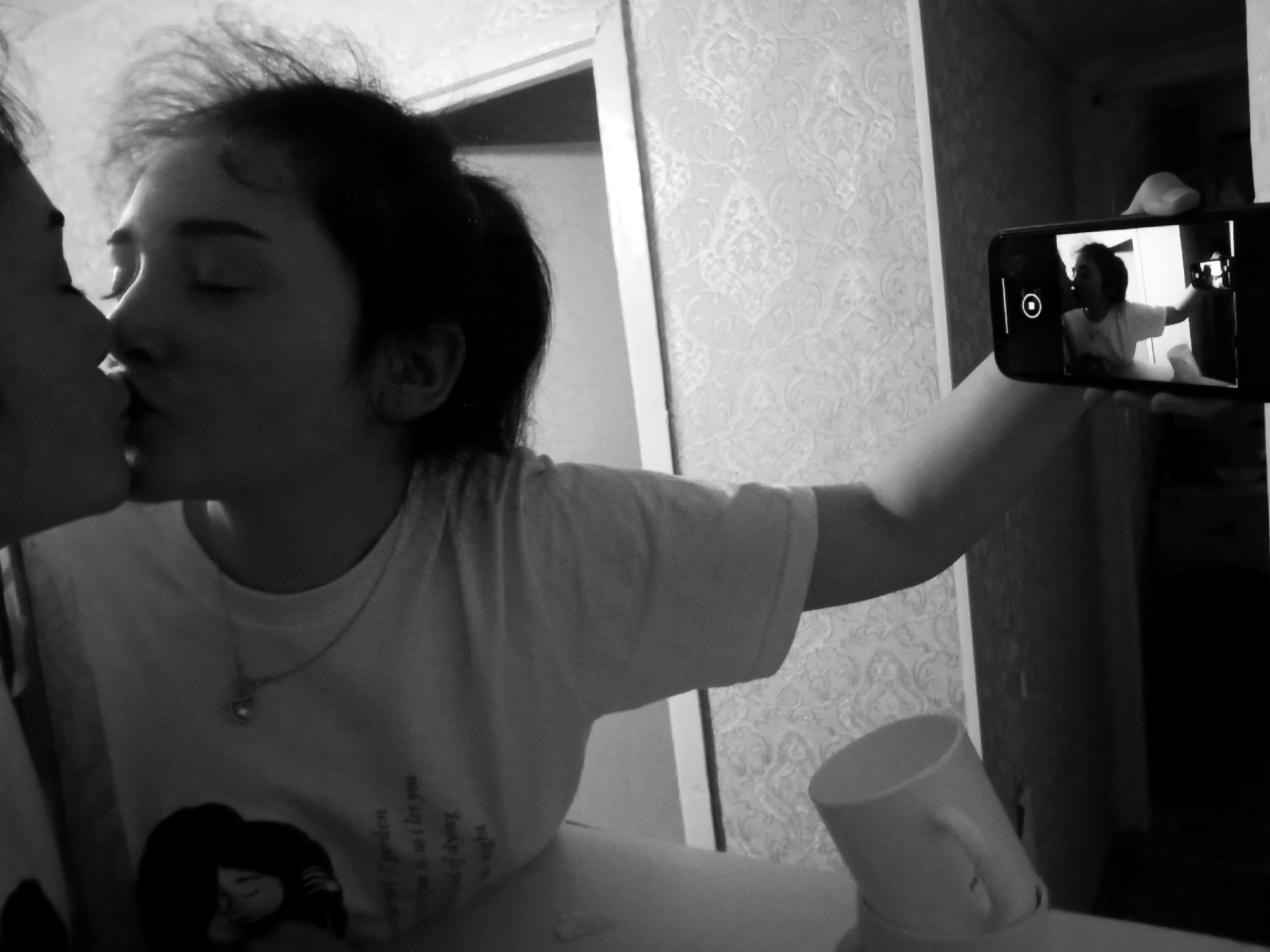 A black and white photograph depicts two individuals in close proximity, potentially kissing. The person on the right holds a smartphone in their outstretched right hand, taking a picture of themselves and the other person. The screen of the phone shows a smaller image of the same scene, creating a mirrored effect. The individual on the right is wearing a t-shirt with a graphic design and a pendant necklace. Their hair is pulled back in a ponytail. A mug or cup is visible in the lower right of the frame. The background includes patterned wallpaper and a doorway. The photograph gives no indication of the city of Poltava, Ukraine.