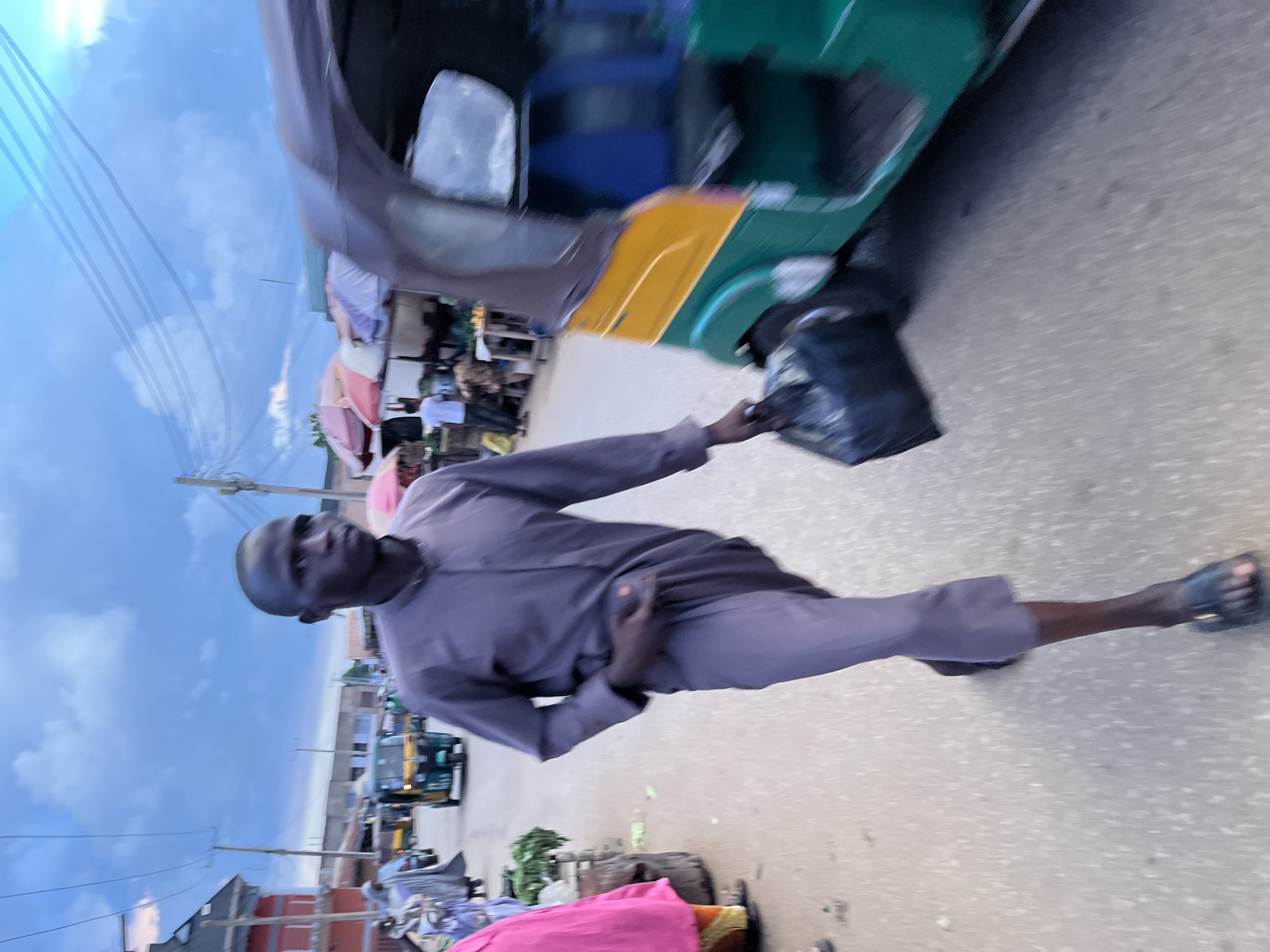 The image presents a motion-blurred, rotated view of an outdoor street scene, likely a market or urban area. The dominant subject is a dark-skinned man with a shaved head, dressed in a loose-fitting, long-sleeved purple or gray tunic and matching pants, wearing dark sandals. He appears to be in motion, extending his right arm to hold a black plastic bag towards a green and yellow auto-rickshaw (tuk-tuk).
The auto-rickshaw is significantly blurred, indicating rapid movement or a moving camera. In the background, other vehicles and structures can be discerned, including buildings and what appear to be market stalls with various colored awnings. A person in a pink garment is partially visible at the bottom edge.
The sky, visible in the upper left, is overcast with distinct clouds and multiple power lines stretching across. This suggests the time of day is either late afternoon, early evening, or a heavily clouded daylight hour, providing diffused light. The ground is a paved surface, likely asphalt or concrete. The entire image has a strong sense of motion and is oriented 90 degrees clockwise from a typical upright perspective. No readable text is visible.
