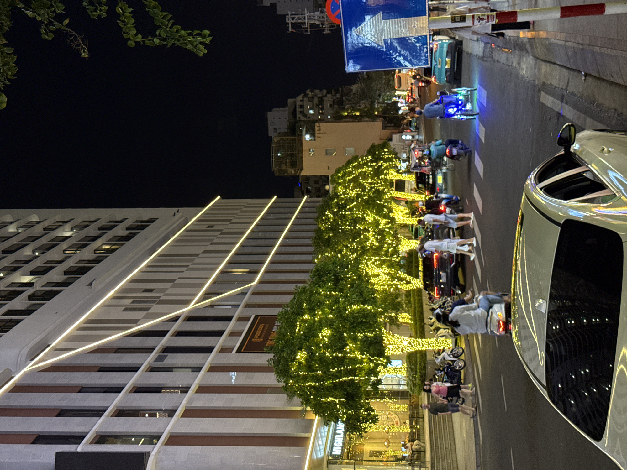 The image captures a street scene at night in Nha Trang, Vietnam, likely along Tran Phu Street, a prominent coastal boulevard. A modern, multi-story building with a facade of vertical cream and reddish-brown panels dominates the left side of the frame. Yellow LED lights are strung diagonally across the building's exterior, creating an illuminated geometric pattern. In front of the building, a row of trees is densely decorated with numerous small, yellow lights, giving them a festive appearance.
The street is moderately busy with traffic. Several motorcycles are visible, with a few individuals riding them and others parked. A white car is in the foreground, partially obscuring the view of the street ahead. Further down the street, a teal-colored car is seen, and in the distance, other vehicles, including what appears to be a bus, are present. A three-wheeled vehicle with blue under-lights is being ridden in the right lane. Pedestrians are walking on the sidewalk on the right, some near a traffic sign with an arrow indicating forward movement. The overall atmosphere is that of a well-lit urban environment at night, decorated for a special occasion or as part of regular street beautification.