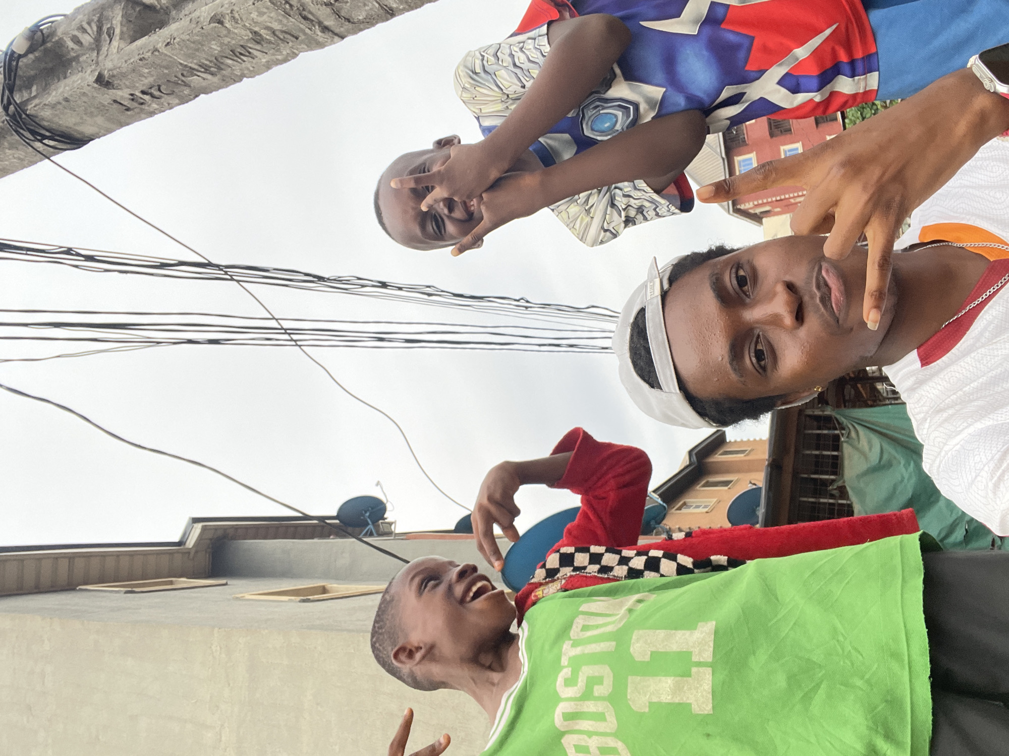 The image shows three Black male youths in an urban area. The location is identified as Onitsha, Nigeria by a sign on a nearby pole "WWUMA 23FT". The youths are standing in close proximity. One youth is wearing a white cap and a red and white shirt. Another youth wears a green shirt with white lettering (partially obscured). The third youth is wearing a blue shirt with light and dark blue pattern on top. The young males are making gestures with their fingers, apparently posing. Overhead power lines are visible against an overcast sky. Buildings are present in the background. There are also satellite dishes on the roofs of the buildings.