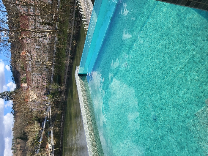 The image displays an elevated outdoor infinity pool, tiled with small turquoise mosaic squares. The pool water reflects patches of white clouds and blue sky. Immediately beyond the pool's infinity edge, a greenish-brown river flows horizontally across the mid-ground. Along the river's far bank, a narrow paved path or road is visible, accompanied by a line of mature deciduous trees largely devoid of leaves. Further back, situated on the riverbank, are several multi-story buildings featuring light-colored facades and distinctive reddish-orange tiled roofs, typical of traditional Portuguese architecture. These structures are nestled amidst additional bare trees. To the far right, a white pedestrian bridge with railings extends along or crosses a section of the river. The background consists of densely wooded hills under a sky with scattered white clouds. No individuals or specific actions are depicted. The scene is located in São Pedro do Sul, Portugal.
