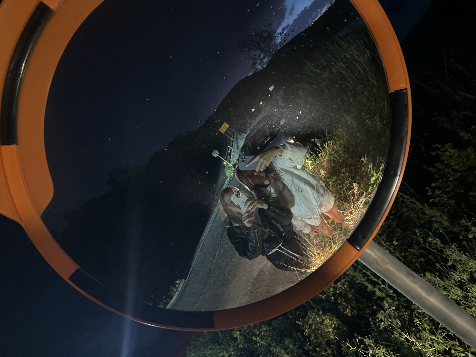 A nighttime photograph captures a distorted reflection in a large, round convex traffic mirror with an orange frame. Two individuals, appearing to be young women, are visible in the reflection, taking a selfie. The person on the right in the reflection wears a light-colored top and holds a light-colored smartphone (likely an iPhone due to the camera array) to capture the image. The person on the left wears a dark jacket, possibly leather.
The reflected background reveals a dark night sky with some visible clouds. Below the sky, a dark, elevated road or path curves into the distance, flanked by dense green and brown foliage and tall grass. A distant, faint light source illuminates a small area on the left side of the reflection, and a yellow, rectangular sign with unreadable text is visible further back. The mirror's support pole is seen on the right, outside the reflective surface, against a dark, vegetated backdrop. The overall scene suggests a casual moment captured late at night in a rural or semi-rural setting in Visnjeva, Montenegro.