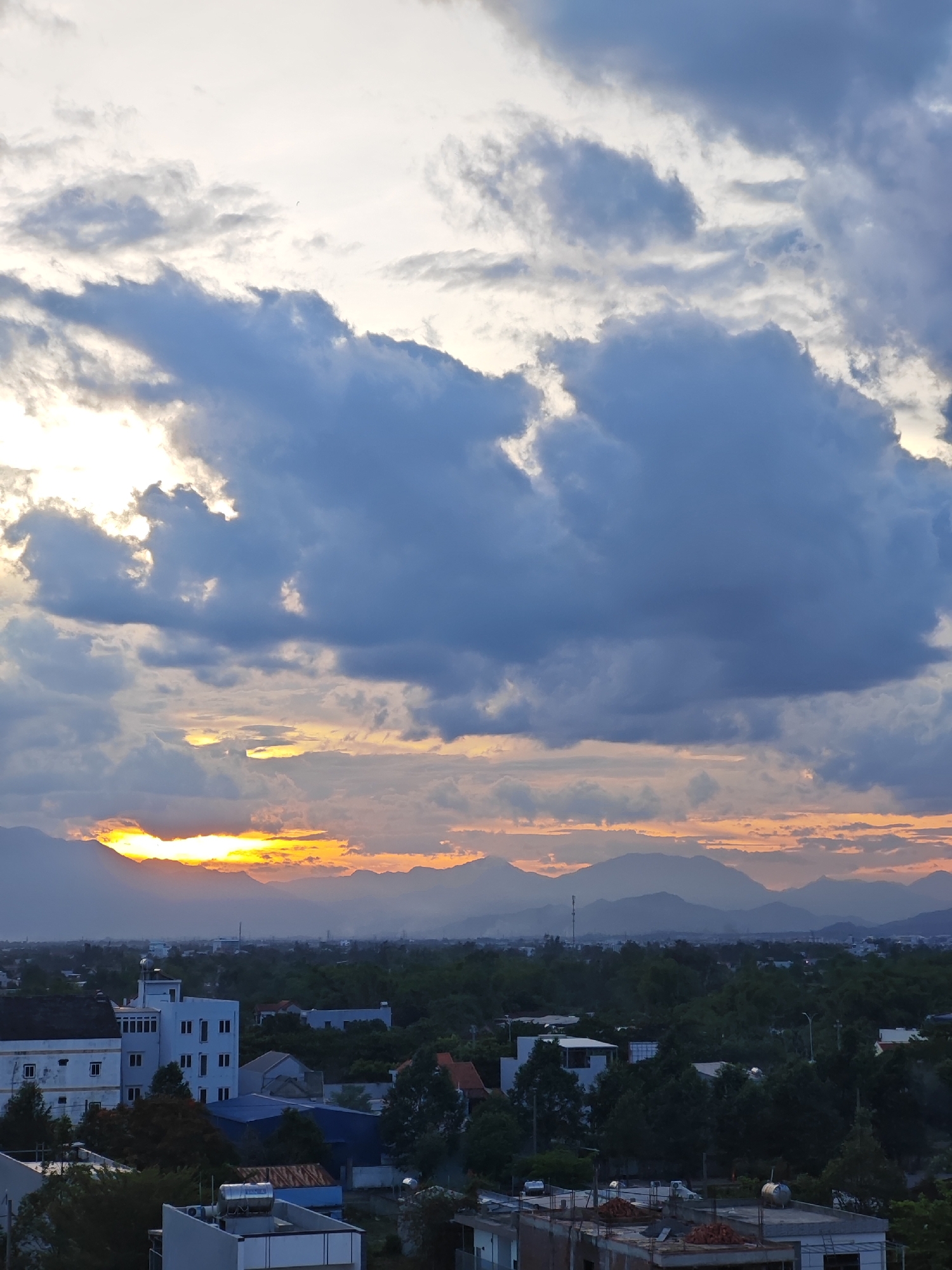 The image captures a panoramic view of the city of Da Nang, Vietnam, during what appears to be sunset or sunrise. The sky is dominated by dramatic, dark clouds, with patches of bright orange and yellow light breaking through, suggesting the sun is low on the horizon and partially obscured. In the foreground, a cluster of buildings and dense green trees are visible. Some buildings appear to be residential or office blocks, with white facades and visible windows. Others are less complete, with exposed construction materials like bricks and concrete. The middle ground features more trees and scattered buildings, interspersed with some infrastructure like a telecommunications tower. In the background, a range of mountains forms a silhouette against the brightly lit sky. The overall scene is one of urban development set against a natural landscape, with the sky providing a striking visual element.