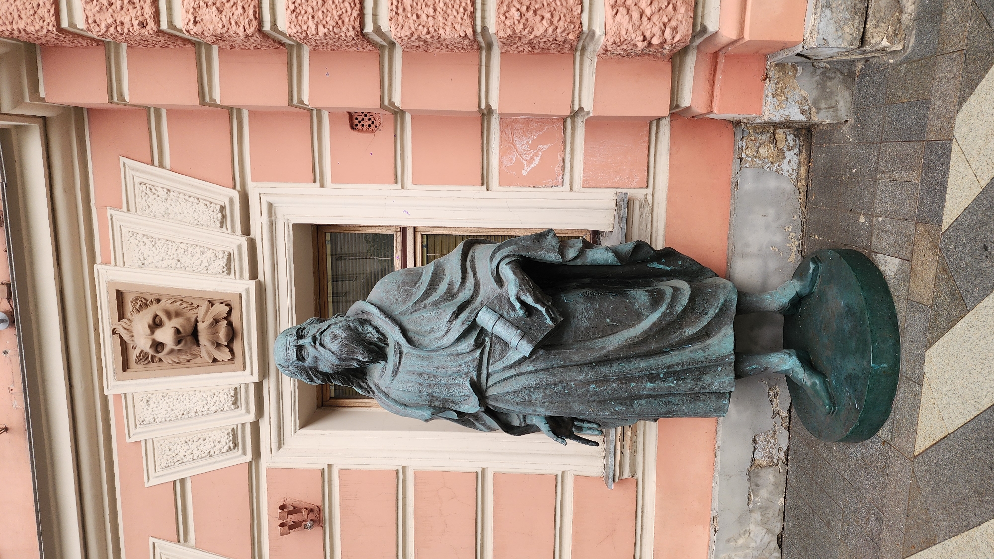 The image shows a bronze statue of a robed figure standing outside a building in Moscow, Russia. The building is painted a salmon pink color with white trim and features decorative elements, including a lion's head relief. The statue is placed on a circular base next to a paved walkway. There's some damage visible on the building's wall near the base of the statue.