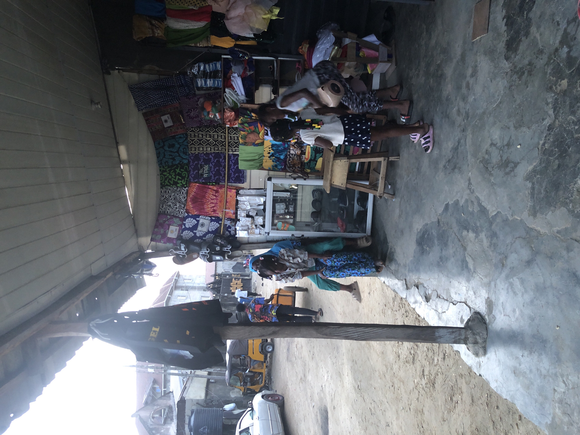 The image depicts an outdoor market scene in a city, possibly in Africa, with stalls displaying various fabrics and goods. In the foreground, a wooden pole supports a canopy, and to the right, a store showcases textiles with vibrant patterns hung on the wall. Two young girls are present: one in a white top and polka dot skirt, and another in a white top and patterned leggings. Behind them, a woman in a blue headscarf and patterned pants sits on a bench. The ground is a mix of dirt and concrete, with a cracked surface. In the background, a white car is parked, and a yellow autorickshaw is visible on the unpaved road. Several buildings are present in the distance. The overall impression is that of a bustling, everyday market.