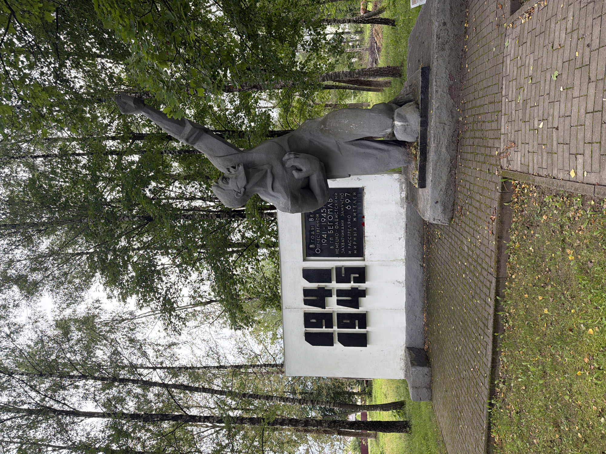 This is a medium shot, eye-level, outdoor photograph of a war memorial located in the Бягомльскі сельскі Савет, Belarus. The memorial consists of a white rectangular monument with large black numbers "1941" and "1945" emblazoned on its left side. To the right of the numbers, a black rectangular plaque contains Cyrillic text. In front of the monument, to the right, is a gray, stone sculpture of a man with his arm raised, his face contorted in anguish or defiance. The sculpture stands on a concrete base. The memorial is situated on a paved walkway surrounded by grass and trees. The trees are tall and slender with green foliage. The ground is covered in green grass with some fallen leaves. The sky is overcast and white. The overall scene is solemn and commemorative.