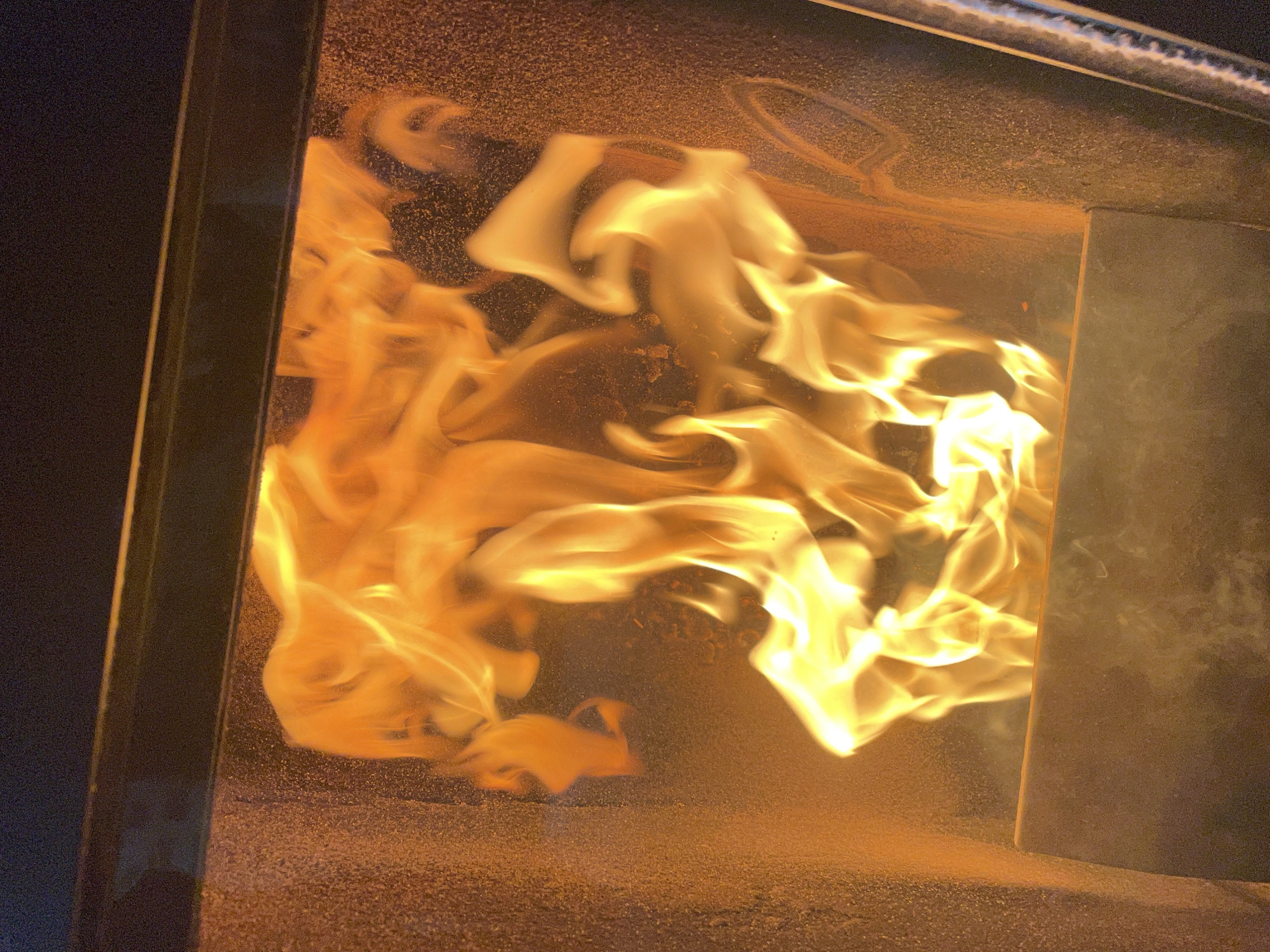 The image depicts a contained fire, possibly within a stove or furnace. The flames are predominantly yellow and white, suggesting intense heat. Ash or particulate matter is visible on the surface below the flames and along the interior walls of the enclosure. The fire is contained within a metallic structure, possibly made of steel or iron. It is not possible to determine if this is located in the city of Zlatograd, Bulgaria from this image.