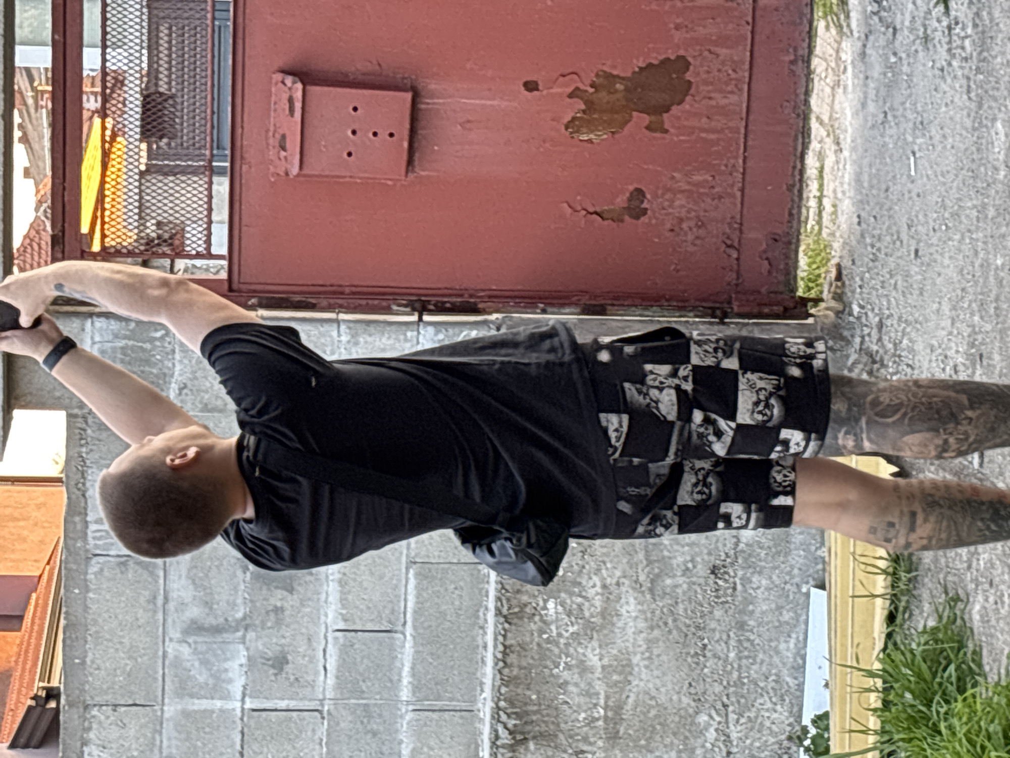 A man with a shaved head is holding a phone in his left hand while standing outside a building. He is wearing a black t-shirt, checkered shorts, and has tattoos on both of his legs. The building has a red door with peeling paint and a rectangular plate with holes. The background also includes a concrete wall, plants, and a yellow metal structure. This scene is in the city of Chirpan, Bulgaria.